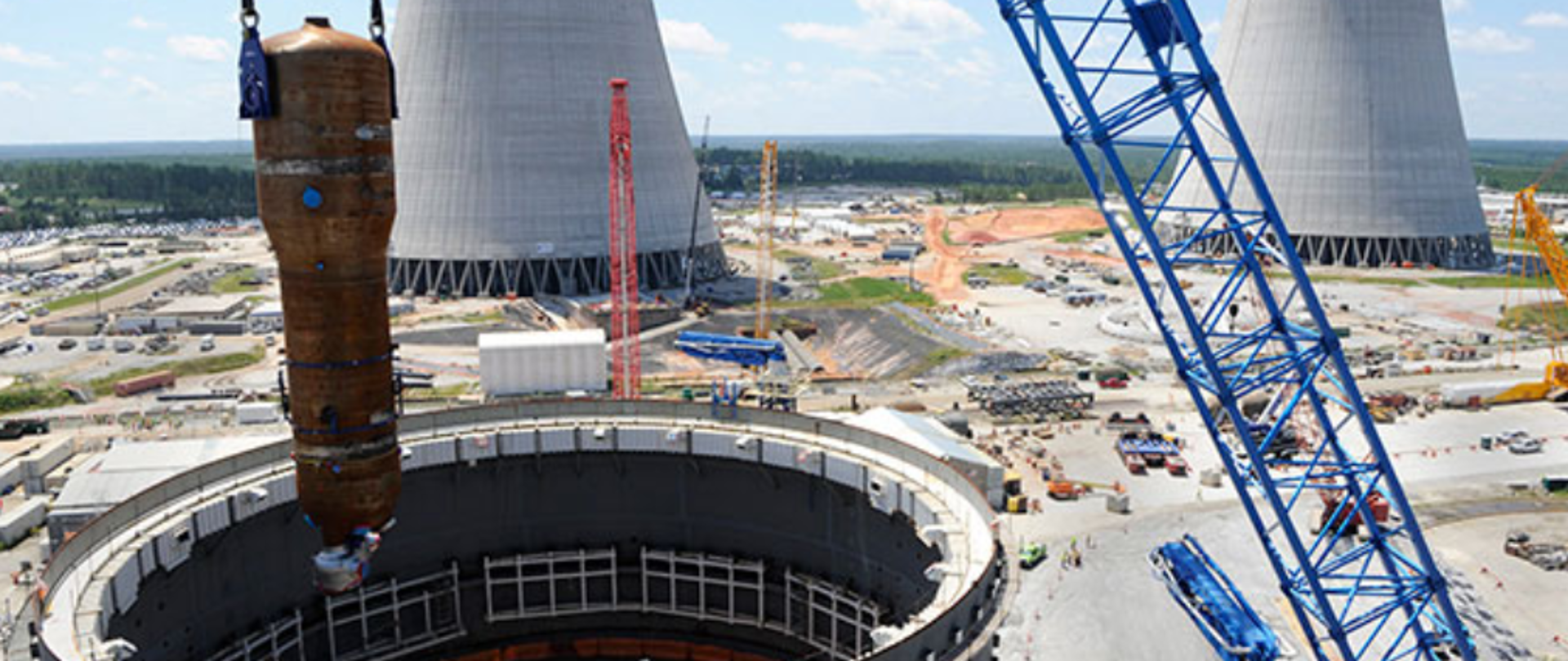 Construction of the Vogtle 3 and 4 Nuclear Power Plant. The blue crane arm places the tank in the nuclear reactor building. Next to the construction site - construction machines. In the background, there are two gray cooling chimneys and a green forest.