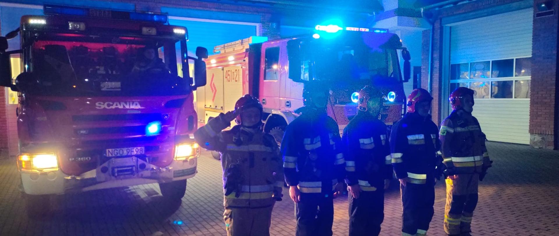 Zdjęcie przedstawia pięciu strażaków Komendy Powiatowej Państwowej Straży Pożarnej w Gołdapi oddających hołd tragicznie zmarłym druhom z OSP Czernikowo, na tle pojazdów pożarniczych z włączonymi sygnałami świetlnymi i dźwiękowymi