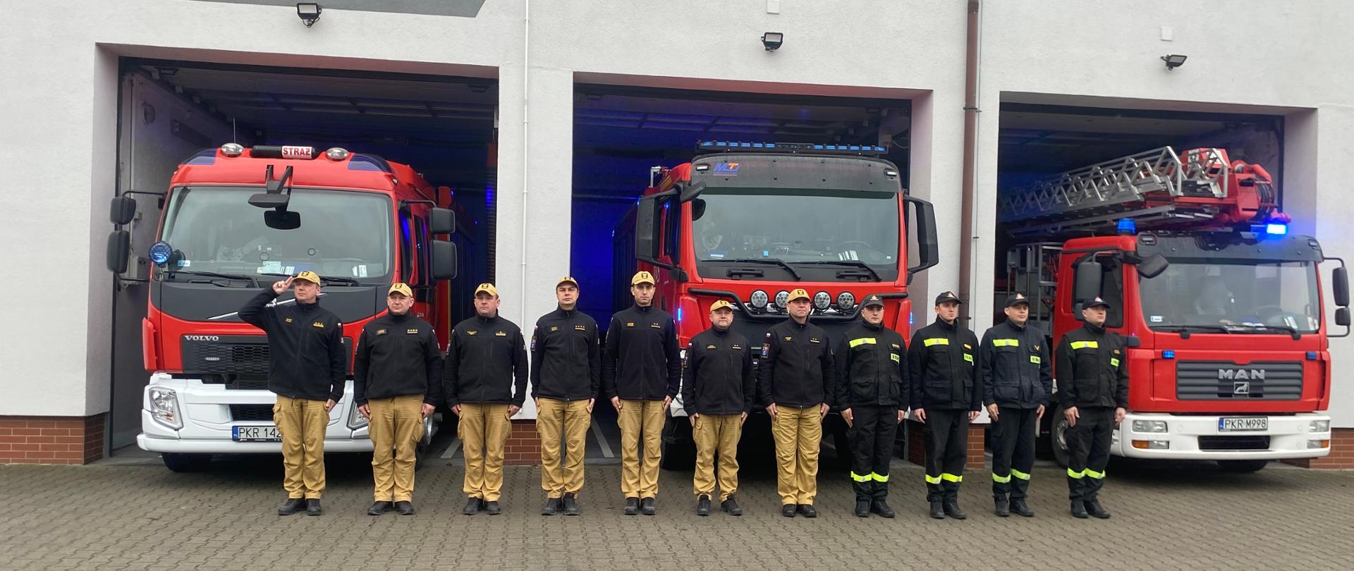 Strażacy stojący w szyku przed pojazdami pożarniczymi, które mają włączone sygnały świetlno - dzwiękowe oddają hołd zmarłemu policjantowi. 