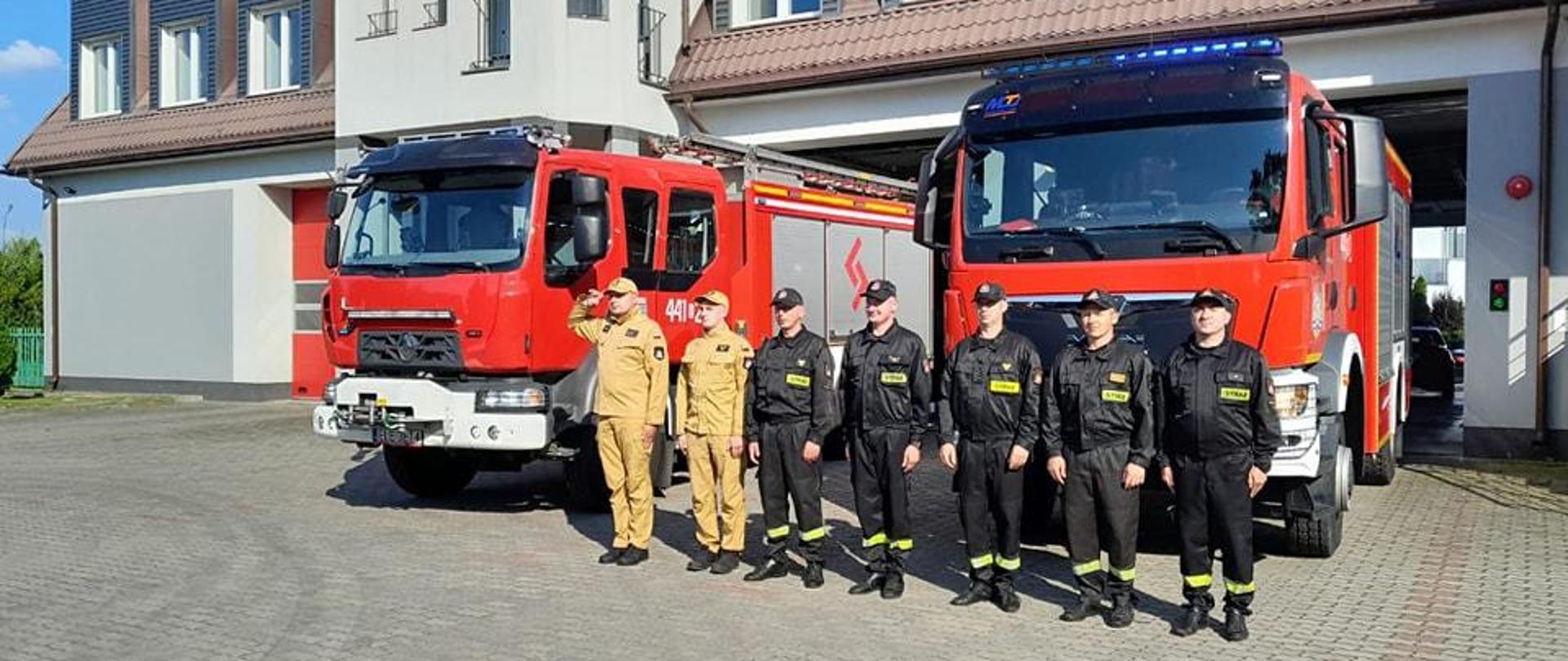 Na zdjęciu widać siedmiu strażaków stojących w jednym szeregu oraz dwa samochody strażackie na placu przed budynkiem Komendy Powiatowej Państwowej Straży Pożarnej w Łęczycy. Pięciu strażaków jest w czarnych ubraniach a dwóch w żółtych. Wszyscy strażacy mają założone czapki z daszkiem. Jest widno.