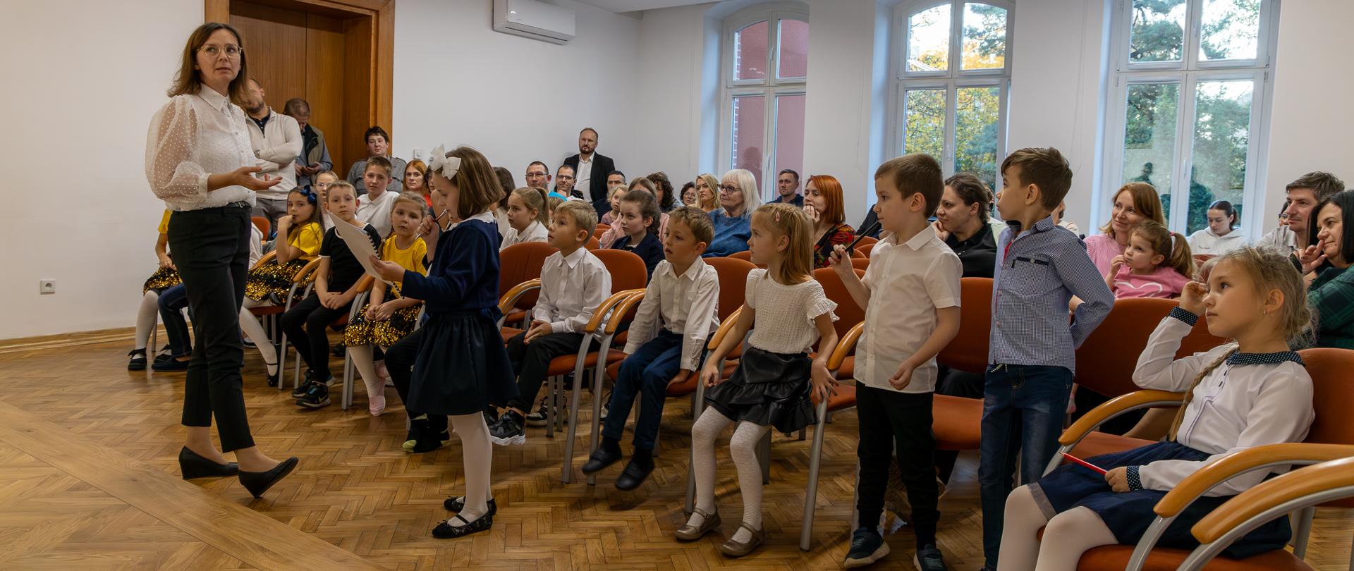 Pasowanie na ucznia. Uczniowie i publiczność zgromadzona na uroczystości podczas rozwiązywania zagadki przygotowanej przez nauczycielki.