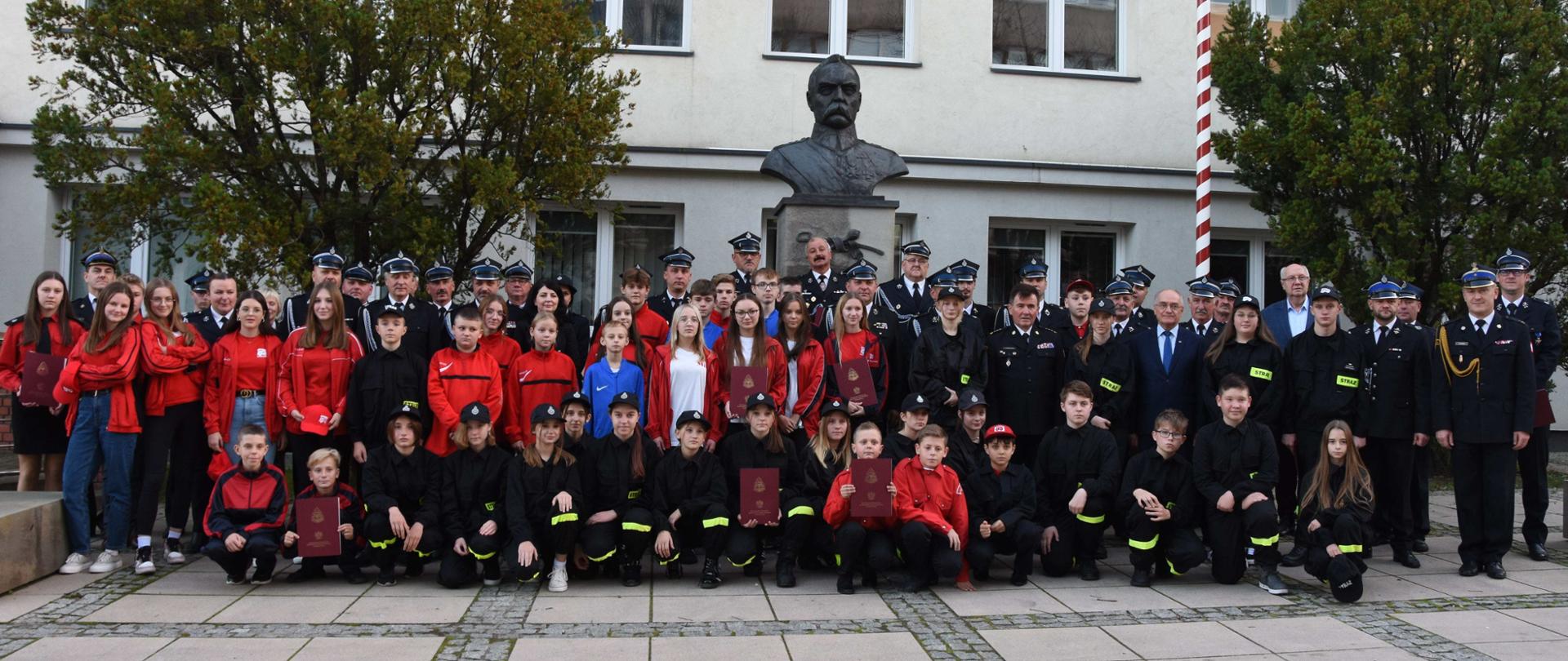 Kolorowa fotografia wykonana w dzień, na zewnątrz na tle jasnej ściany budynku z wieloma oknami. Przed budynkiem stoi grupa około 70 osób dorosłych oraz młodzieży z dyplomami. Dorośli są w mundurach strażaków ochotników i funkcjonariuszy Państwowej Straży Pożarnej. Przed dorosłymi licznie zgromadzone dzieci są mundurkach Młodzieżowych Drużyn Pożarniczych. Za stojącymi widoczny pomnik z popiersiem, biało-czerwony maszt flagowy i dwa zielone krzewy.