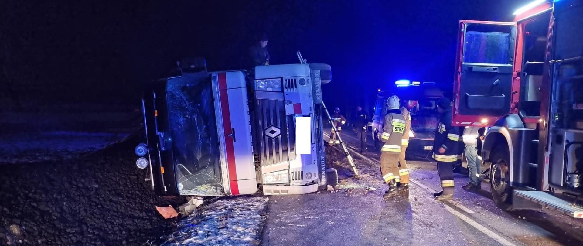 Zdjęcie przedstawia strażaków prowadzących działania w obrębie pojazdu ciężarowego. Widoczne są także pojazdy Straży Pożarnej