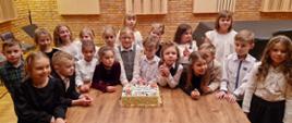Grupa dzieci stoi przy stoliku. Na stoliku widoczny kolorowy prostokątny tort z napisem "Gratulujemy pierwszego występu w Filharmonii"