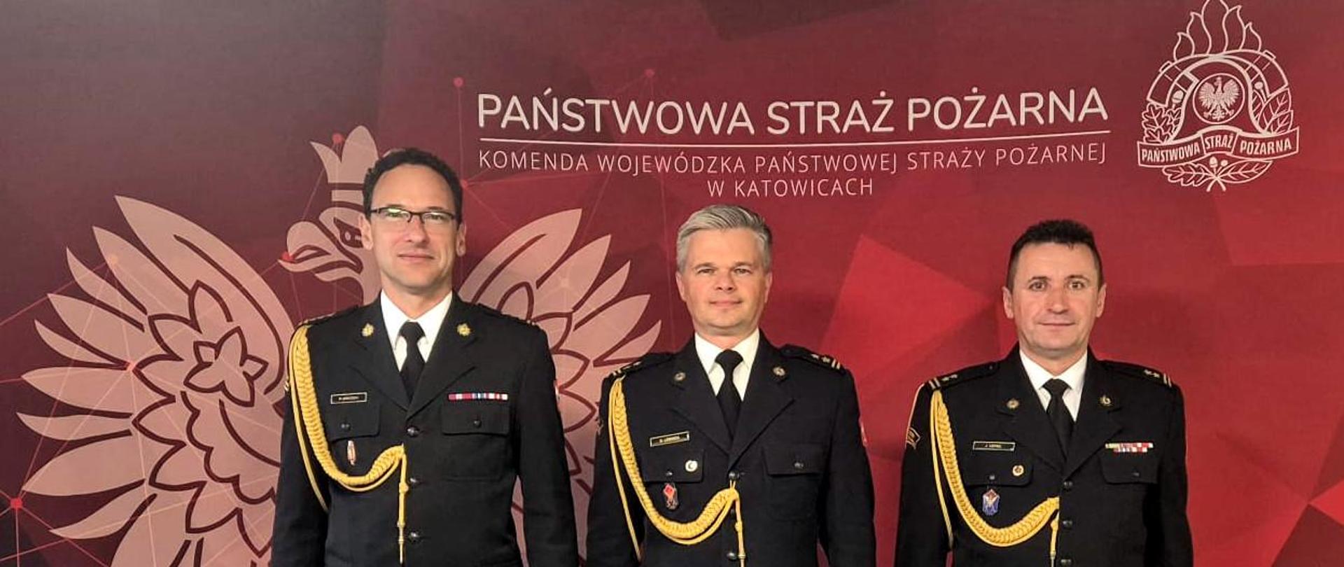 Na tle czerwonej ścianki z białym orłem w koronie po lewej stronie i logo Państwowej Straży Pożarnej po prawej. Na środku napis Państwowa Straż Pożarna.Komenda Wojewódzka PSP w Katowicach, stoją w mundurach wyjściowych, od prawej śląski komendant wojewódzki st. bryg. Wojciech Kruczek, w środku nowo połowany zastęcpca bryg. Daniel Lebioda i komendant miejski PSP w Jastrzębiu Zdroju bryg. Jan Cepiel