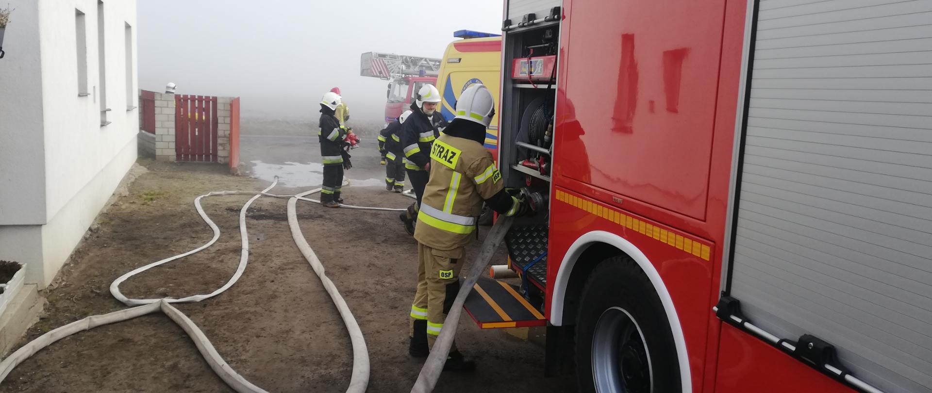 Na zdjęciu po prawej stronie widać czerwony samochód strażacki, który stoi na podwórku prywatnej posesji. Przy nim stoi strażak, który odkręca wąż pożarniczy. Bezpośrednio za wozem strażackim widać tył karetki pogotowia a przy niej kilku strażaków zwija węże pożarnicze. W tle widać również przód drabiny mechanicznej. Po lewej stronie zdjęcia widać szczyt budynku mieszkalnego