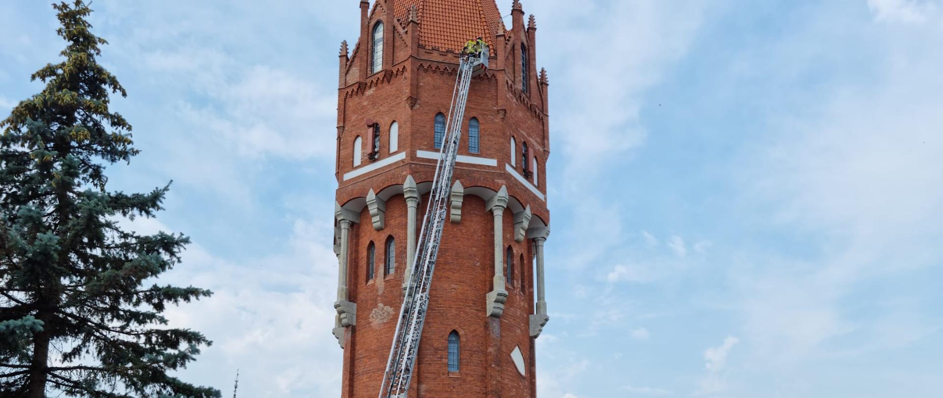 Fotografia przedstawia strażaków w koszu drabiny mechanicznej wysuniętej w górę wieży ciśnień na wysokość 37 metrów