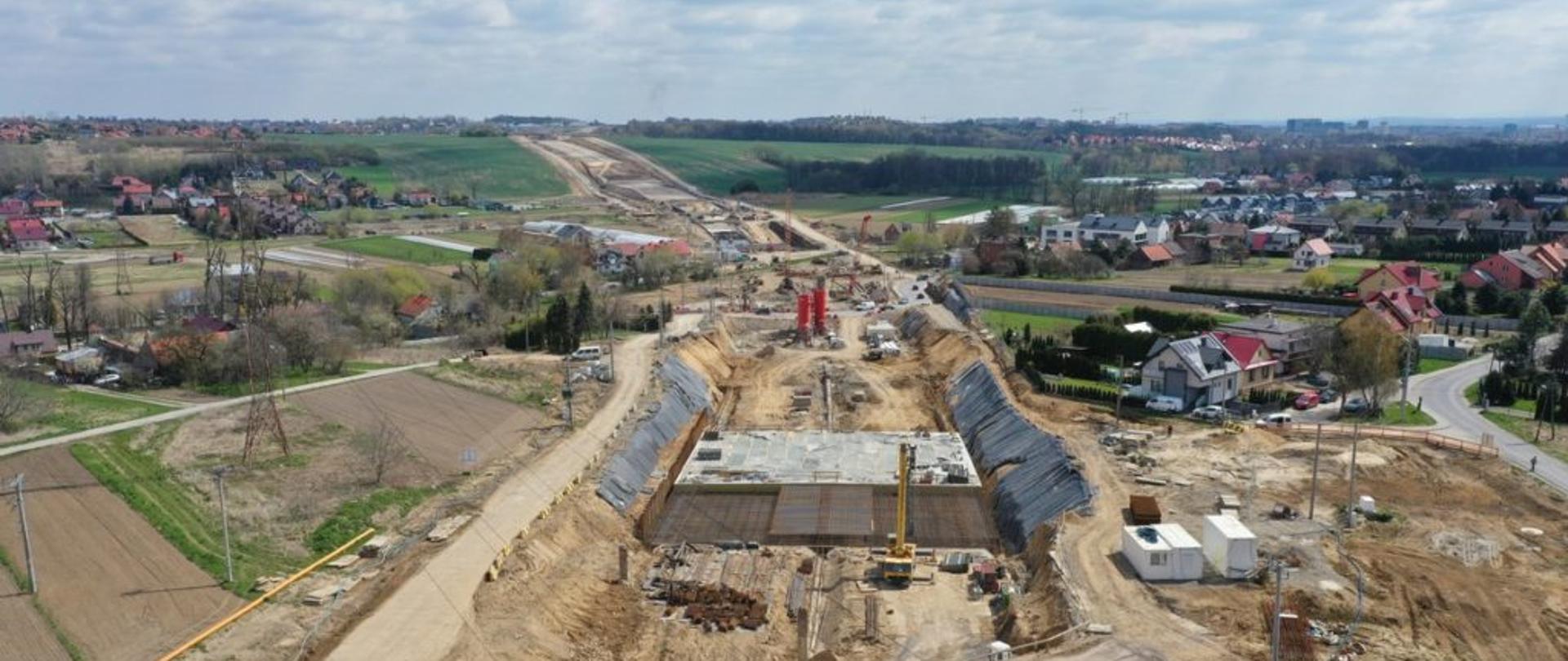 Zdjęcie przedstawia plac budowy drogi ekspresowej S52, znanej również jako Północna Obwodnica Krakowa. Na pierwszym planie widoczny jest szeroki wykop, w którym trwają prace budowlane. W centralnej części zdjęcia znajduje się betonowa konstrukcja. Wokół placu budowy widać różne maszyny budowlane, takie jak dźwigi i koparki. Po obu stronach wykopu znajdują się wzmocnienia skarp, wykonane z metalowych siatek. W tle rozciąga się widok na okoliczne tereny, w tym pola uprawne, domy mieszkalne oraz dalsze etapy budowy drogi. Na horyzoncie widać zalesione wzgórza oraz zabudowania miejskie.