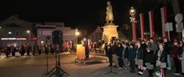 Plac przed Pomnikiem Żołnierza Polskiego w Rawiczu. W centralnym punkcie znajduje sę pomnik stojący na postumencie. Wokół niego zgromadzeni sa uczestnicy uroczystości Na podwyższeniu, przy mównicy stoi strażak prowadzący apel. W tle budynki. 