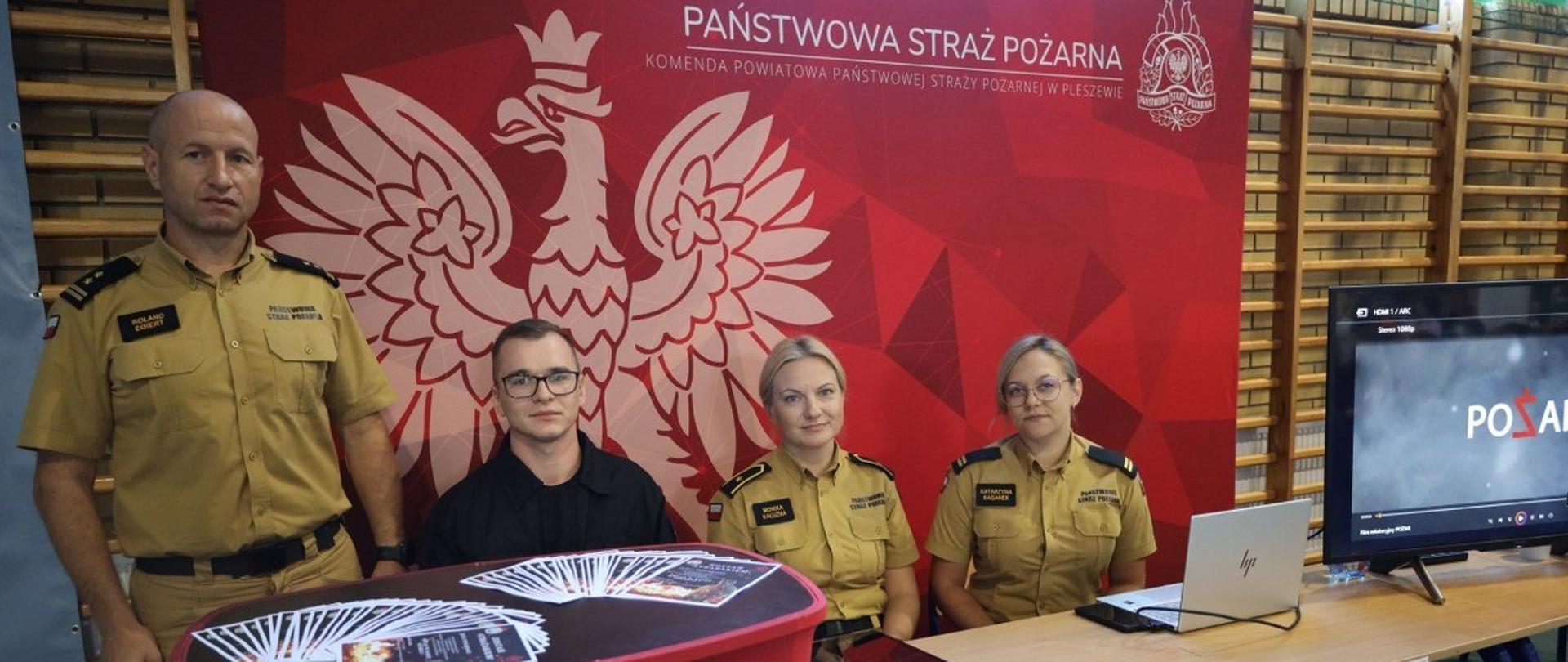 Przed czerwonym banerem z napisem Państwowa Straż Pożarna siedzą dwie kobiety w żółtych mundurach Państwowej Straży Pożarnej i mężczyzna w czarnym mundurze Państwowej Straży Pożarnej. Po lewje stronie za mównicą stoi mężczyzna w żółtym mundurze Państwowej Straży Pożarnej, na stoliku przed nimi leżą ulotki i stoi telewizor.