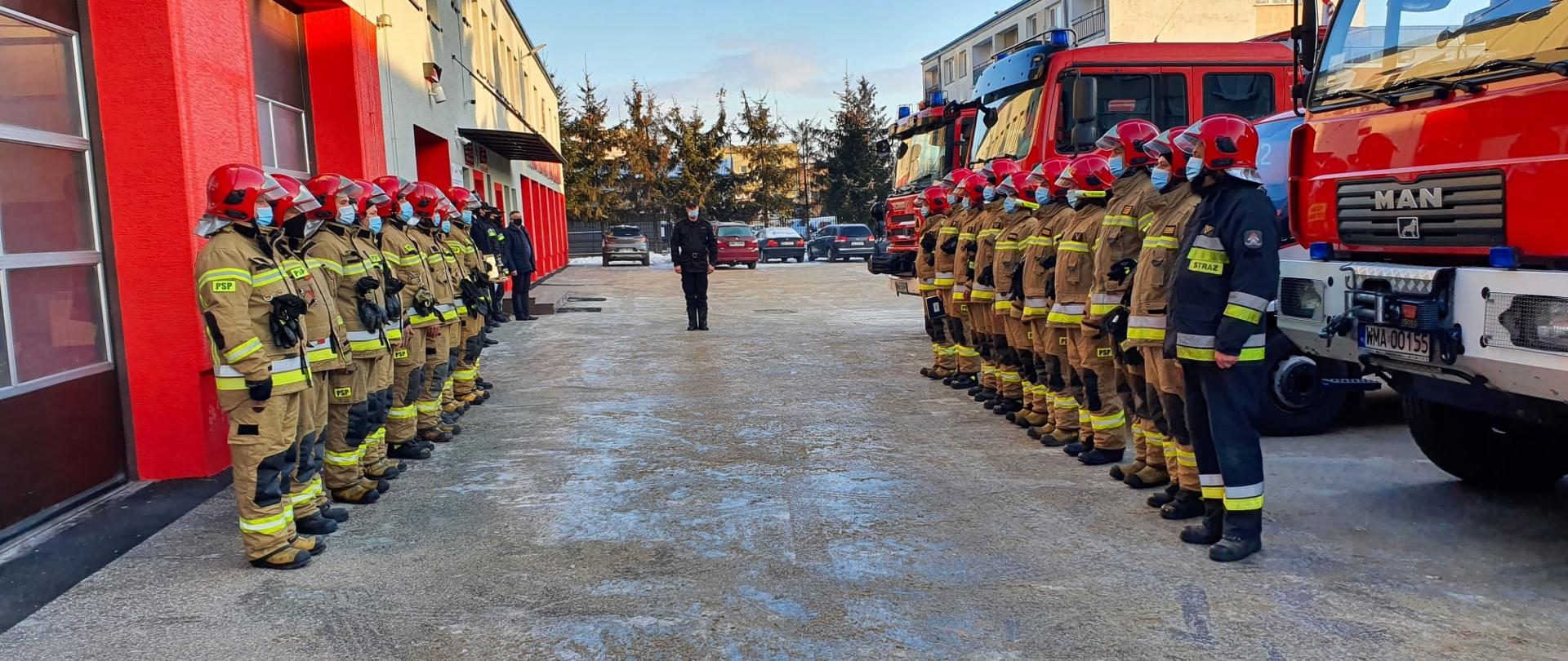 Uroczysta zmiana służby na placu wewnętrznym Komendy. Dwie zmiany służbowe w ubraniach specjalnych stoją w szeregu naprzeciwko siebie. Jedni mają za plecami budynek Komendy, drudzy samochody gaśnicze. Po środku między nimi stoi Dowódca JRG.