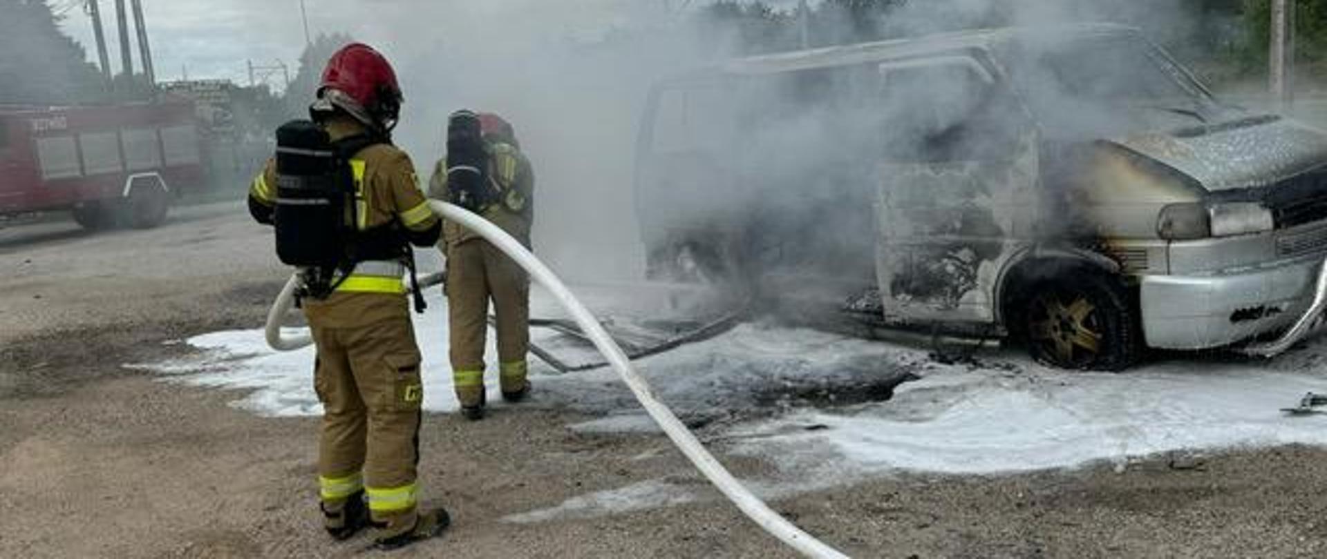 Zdjęcie przedstawia dwóch ratowników podczas akcji gaśniczej. Przed nimi stoi spalony całkowicie dostawczy Volkswagen.