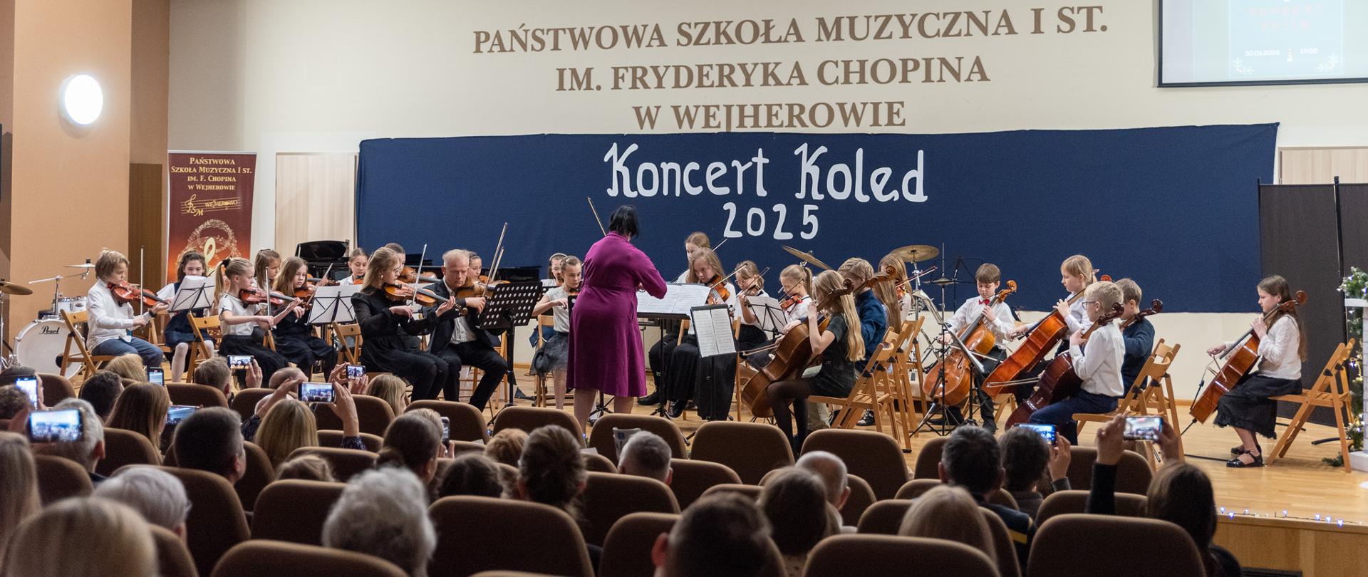 Wystąp orkiestry smyczkowej podczas Koncertu Kolęd. Dyryguje Agata Parzych. Na pierwszym planie widoczna liczna publiczność.