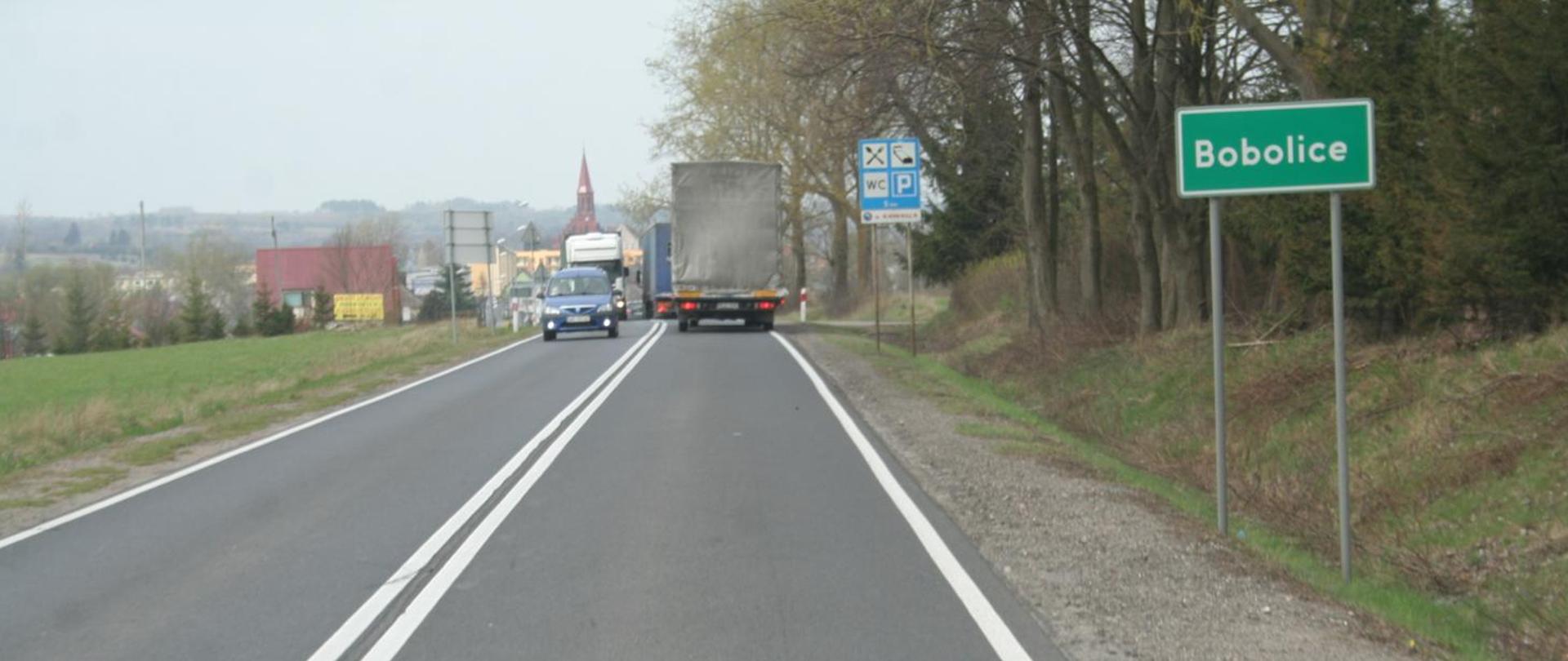 Pojazdy poruszające się drogą krajową. W tle widoczne zabudowania, a po prawej stronie znak, informujący o wjeździe do miejscowości Bobolice.