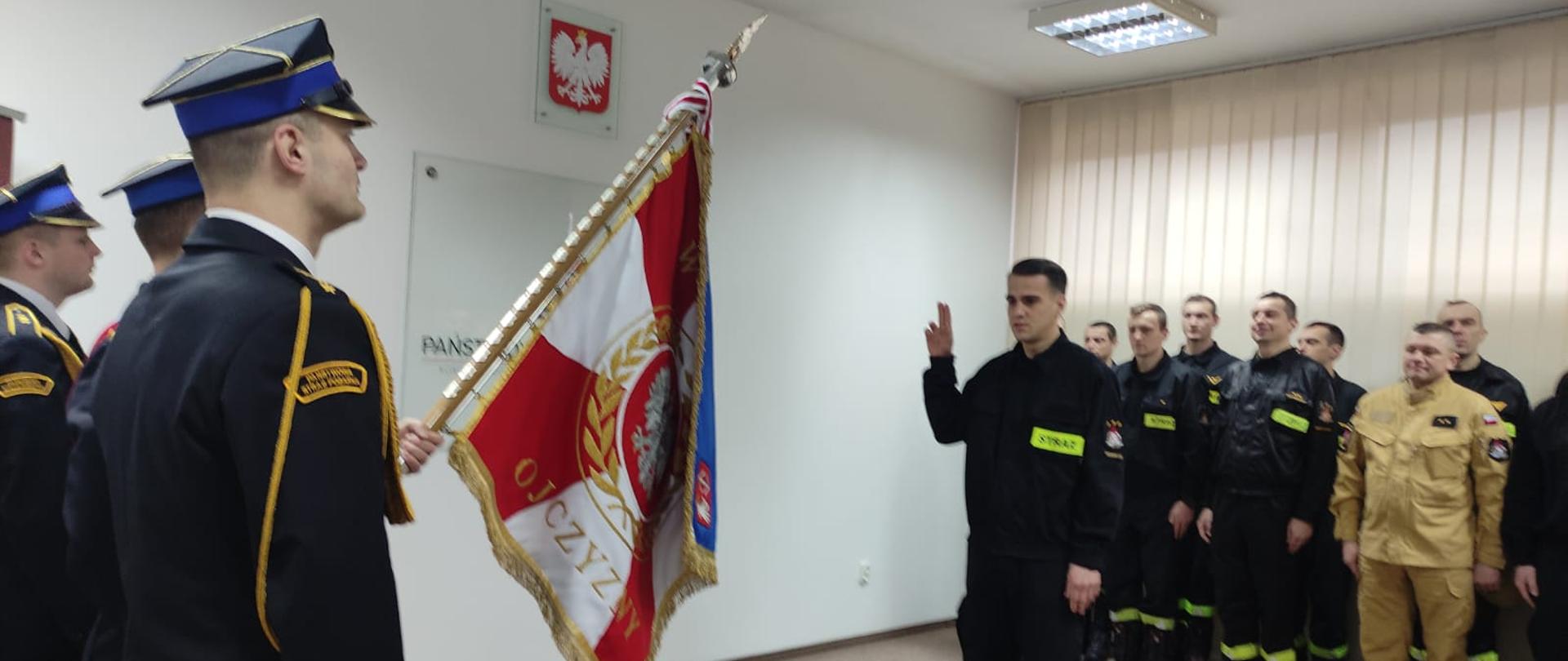 Str. Sebastian Wyleziński składa przed sztandarem KP PSP Grodzisk Maz. ślubowanie. tyłu za nim stoją w dwuszeregu strażacy ze zmiany przyjmującej i zdającej.