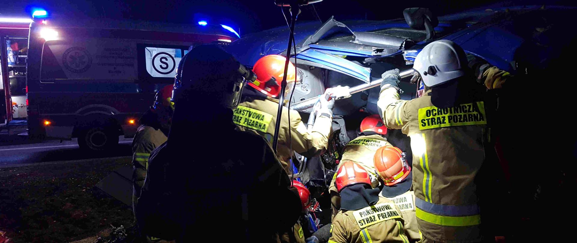 Zdjęcie zrobione w nocy. Na zdjęciu widać strażaków jak przy pomocy narzędzi hydraulicznych wycinają zakleszczonego kierowcę z samochodu ciężarowego. W dalszej części zdjęcia widać karetkę pogotowia ratunkowego i samochód strażacki. Jeden strażak oświetla teren działań przy pomocy najaśnicy. 