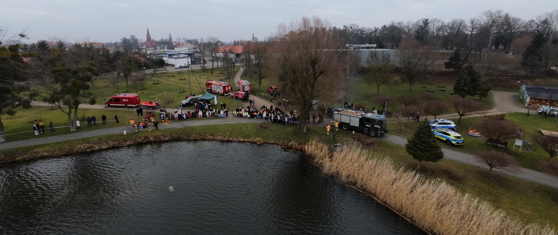 widok znad jeziora, grupa ludzi stoi przy brzegu akwenu wodnego, strażacy stoją przy lustrze wody, w tle samochody strażackie