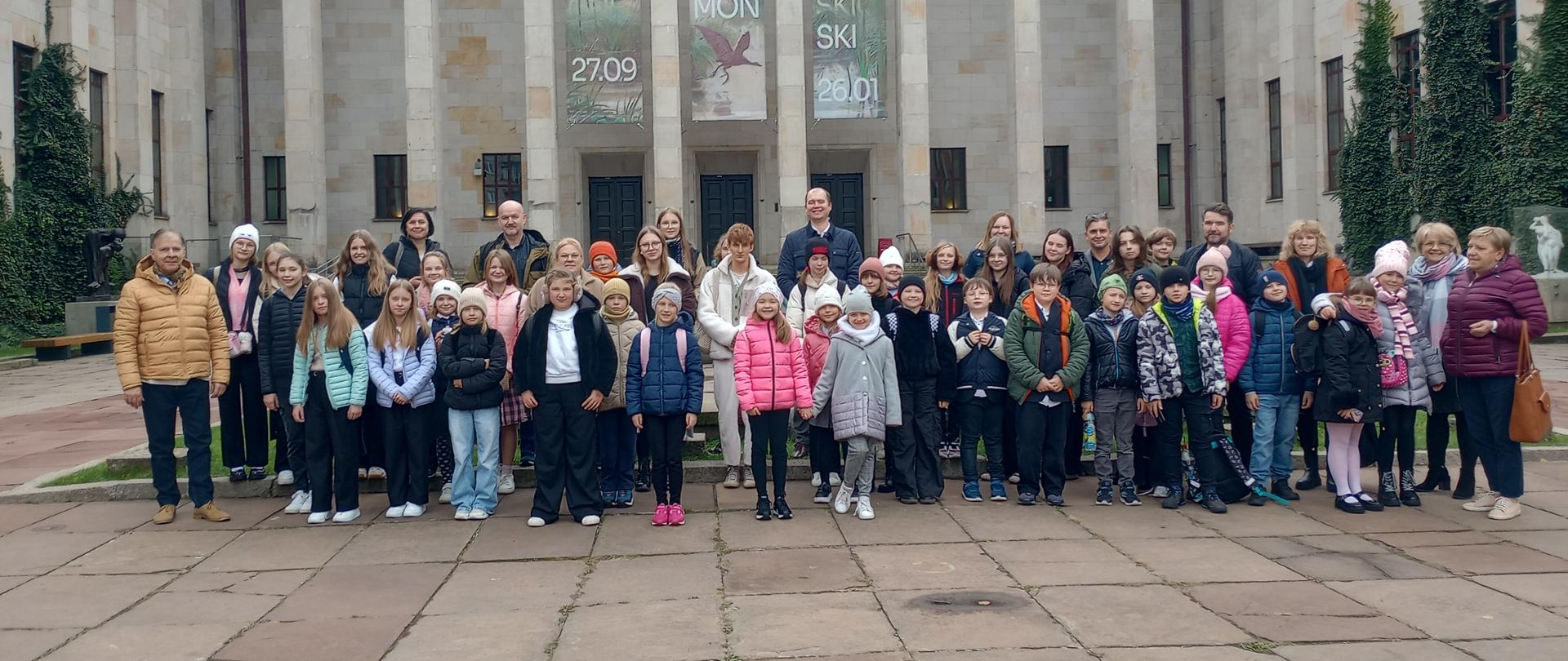 Zdjęcie zbiorowe 50-osobowej grupy dzieci, młodzieży, dorosłych. Wszyscy ustawieni są na tle budynku Muzeum Narodowe w Warszawie. Wszyscy są uśmiechnięci i ciepło ubrani.