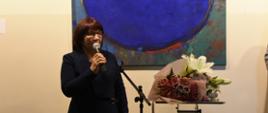 Kobieta stoi z mikrofonem w ręku, w tle obraz, obok kwiaty