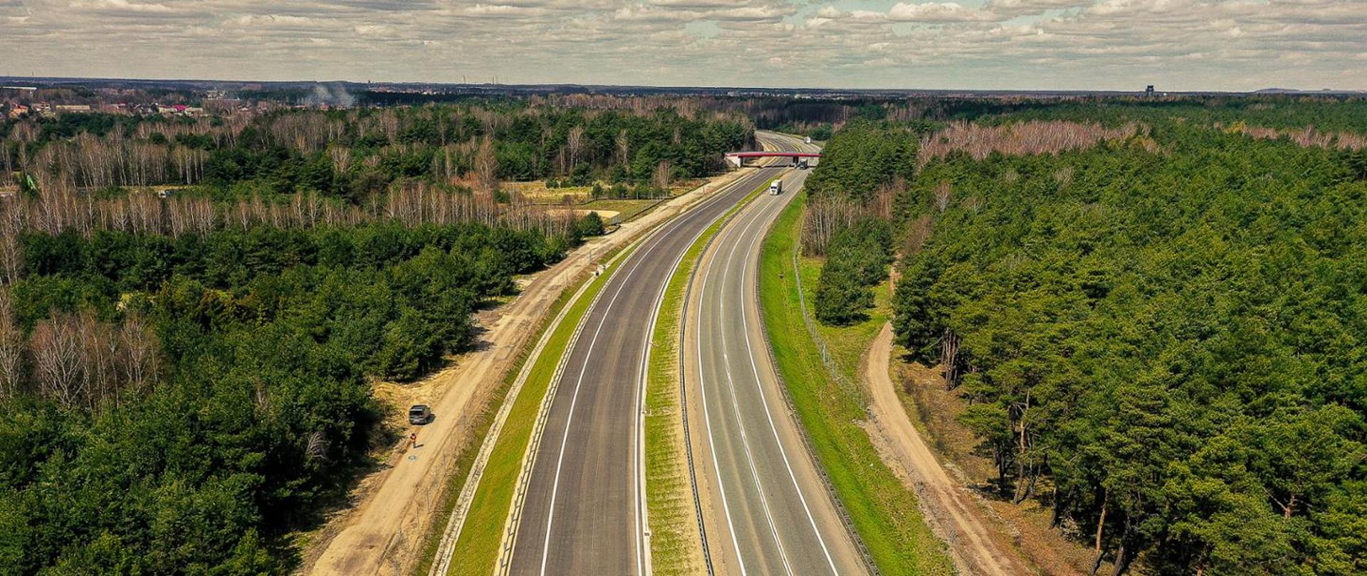 Fotografia lotnicza dwujezdniowej drogi, o dwóch pasach ruchu w każdym z kierunków. Droga przebiega przez tereny zalesione. Poniżej horyzontu drogę przecina poprzecznie wiadukt z czerwonymi belkami gzymsowymi.