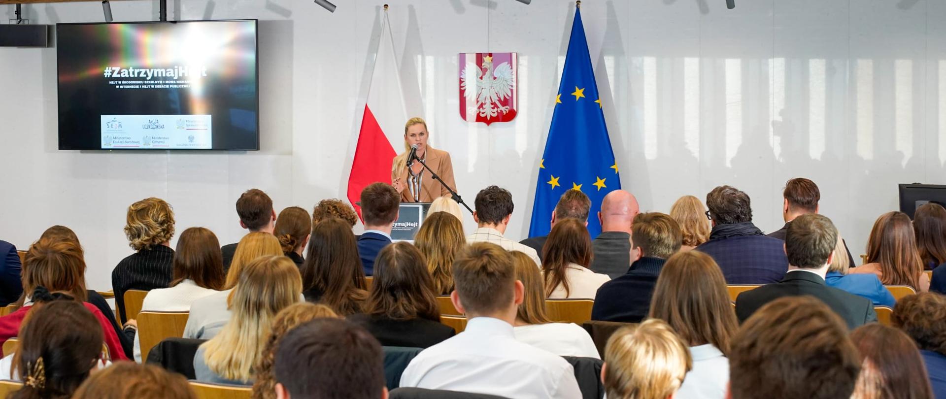 Widok z tyłu na salę. Minister Nowacka stoi przy pulpicie i przemawia do mikrofonu. Za nią dwie flagi Polski i UE oraz po lewej stronie duży telewizor na którym wyświetlany jest tekst #ZatrzymajHejt. Przed minister sala wypełniona ludźmi.