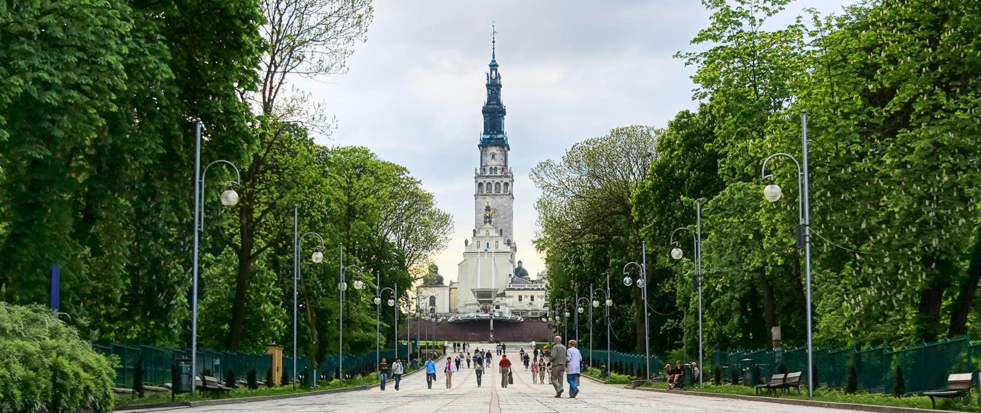 Po prawej i lewej stronie ukazane są zielone drzewa, pośrodku spacerują ludzie. Na wprost znajduje się bazylika