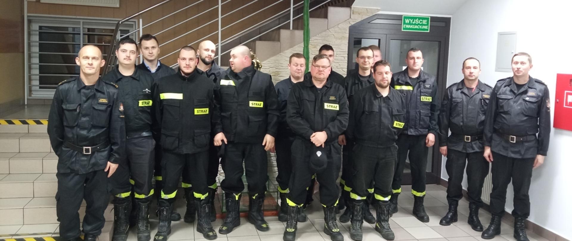 zdjęciu strażacy z Ochotniczych Straży Pożarnych po egzaminie kończącym szkolenie naczelników OSP. Strażacy ubrani w ubrania koszarowe koloru czarnego bez czapek na holu Komendy Powiatowej Państwowej Straży Pożarnej w Gorlicach.