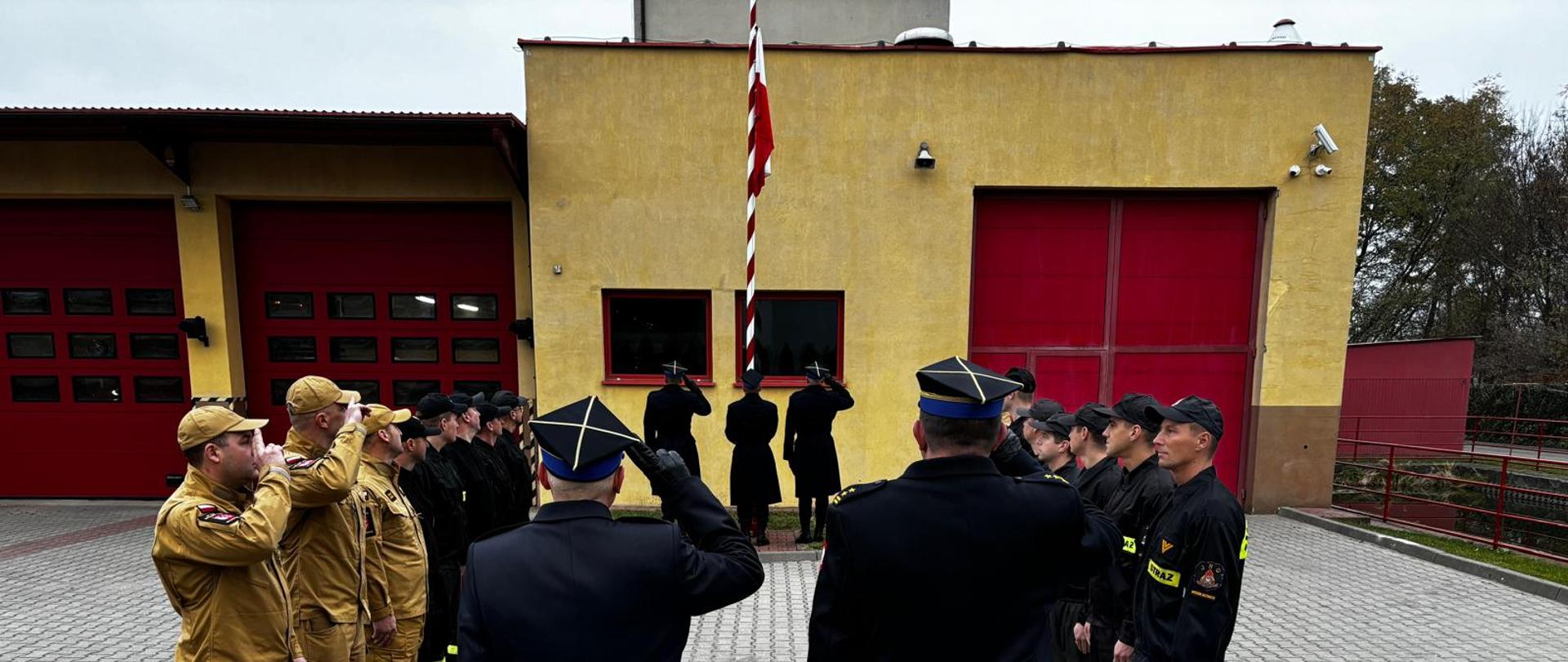 Strażacy oddają honory fladze państwowej. Poczet flagowy stoi przed masztem flagowym jeden ze strażaków z pocztu wciąga flagę na maszt. Zmiana zdająca jest po lewej stronie. Zmiana przyjmująca jest po prawej stronie. Komendant Powiatowy PSP i Dowódca JRG są na środku