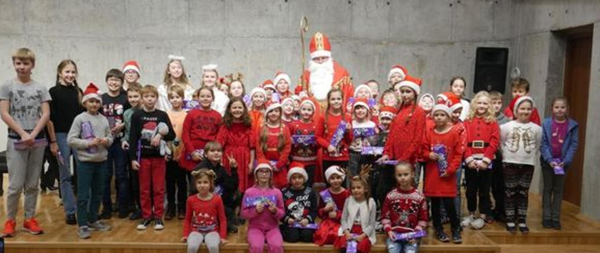 Zdjęcie przedstawia grupę dzieci w czerwonych czapkach siedzących na scenie wokół Świętego Mikołaja
