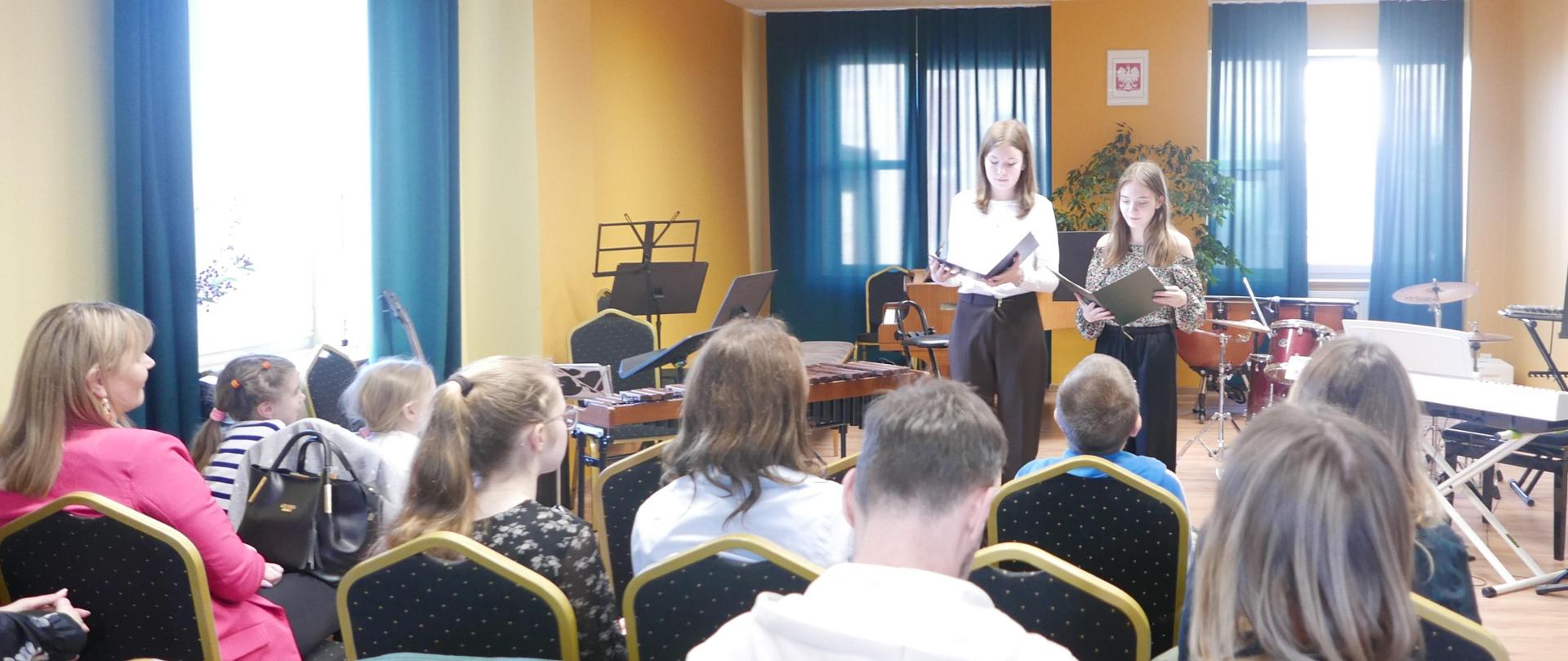 Aula szkoły muzycznej, publiczność . Na scenie dwie uczennice szkoły zapowiadające koncert.