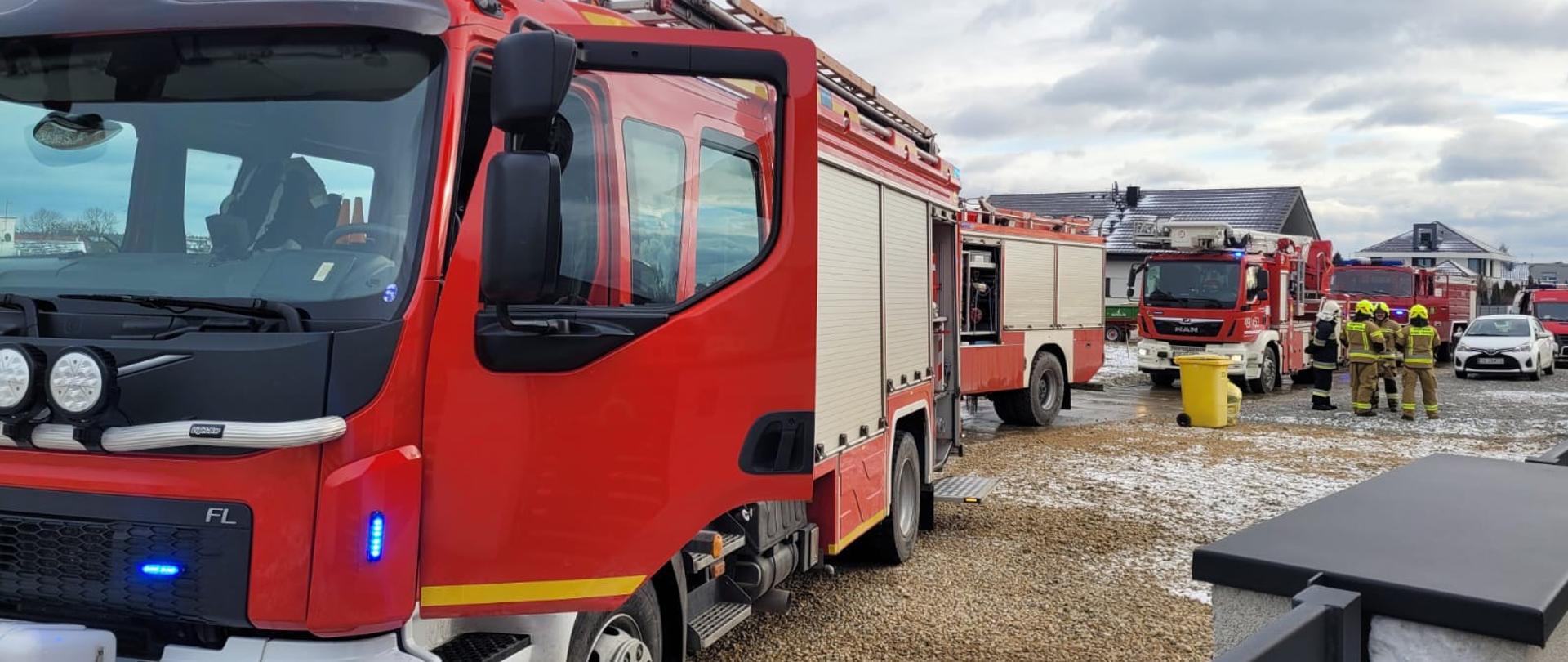 Na zdjęciu znajdują się samochody strażackie, strażacy w nomexach, samochód osobowy i budynki mieszkalne