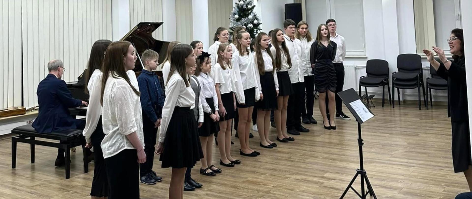 Koncert kolęd. W sali koncertowej śpiewa chór złożony z uczennic i uczniów PSM I st. w Pułtusku. Przed nimi stoi nauczycielka szkoły i dyryguje. Za nimi siedzi dyrektor szkoły i akompaniuje na fortepianie. 