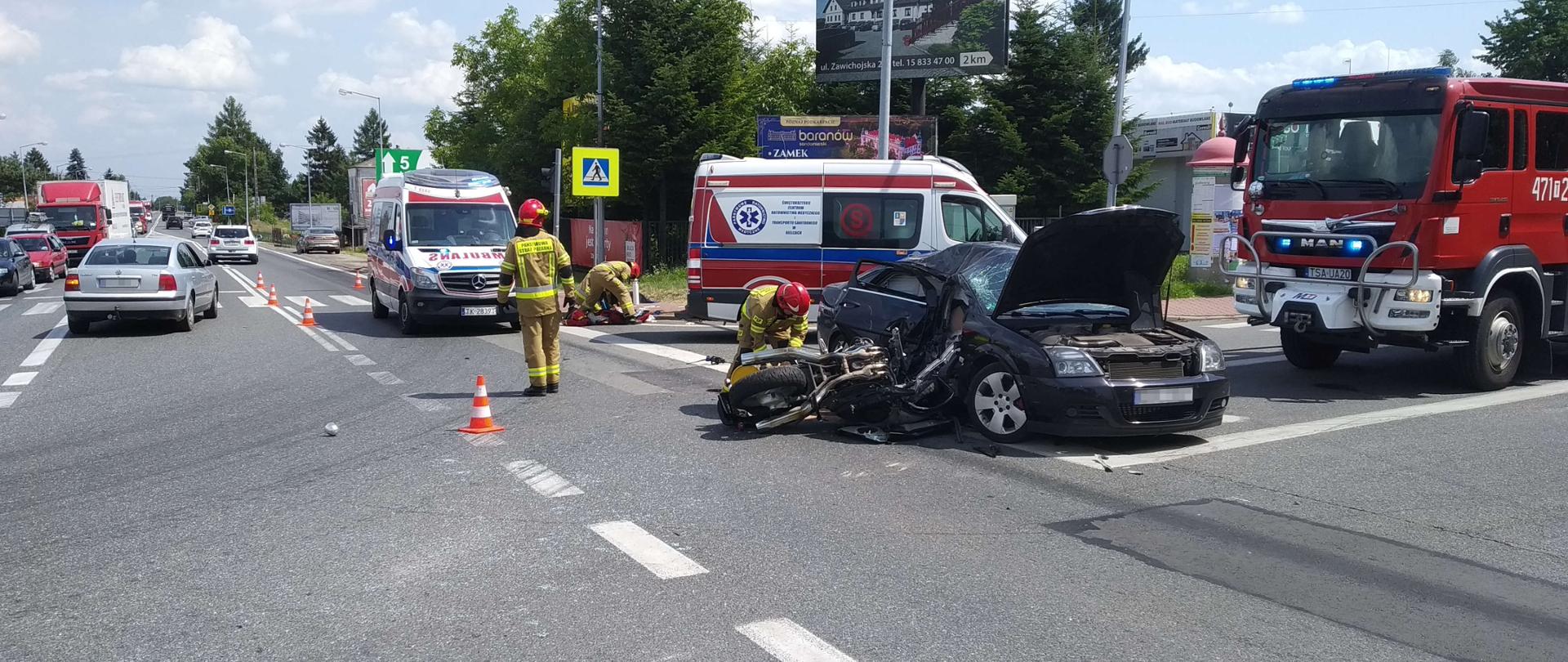 Zdjęcie zrobione w dzień. Zdjęcie przedstawia skrzyżowanie ulic. Na pierwszym planie widać rozbity samochód osobowy oraz motocykl, przy których strażak prowadzi działania. W tle widać strażaka udzielającego KPP poszkodowanemu pomiędzy dwiema karetkami.