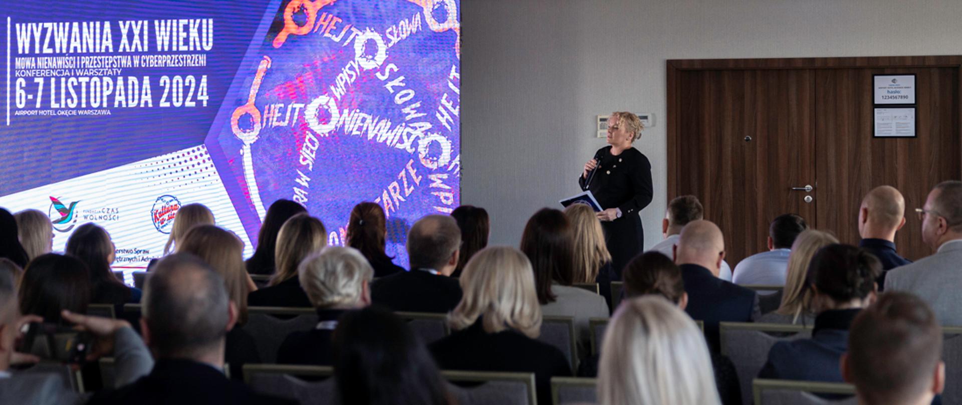 Dyrektor Agata Furgała otwiera spotkanie - przemówienie na tle ścianki z nazwą konferencji : Konferencja i warsztaty "Wyzwania XXI wieku - mowa nienawiści i przestępstwa w cyberprzestrzeni"