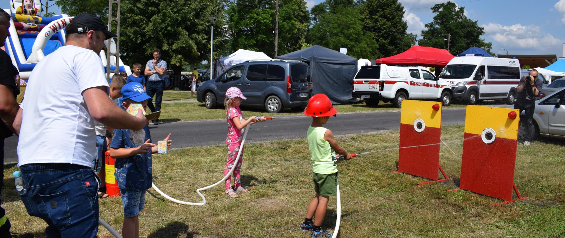 Zdjęcie przedstawia dwójkę dzieci ( chłopca i dziewczynkę ) trzymających małe węże z wodą i celujące w tablicę. Dookoła widać samochody oraz namioty z rozstawionymi stoiskami, dmuchańce do zjeżdżania oraz ludzi.