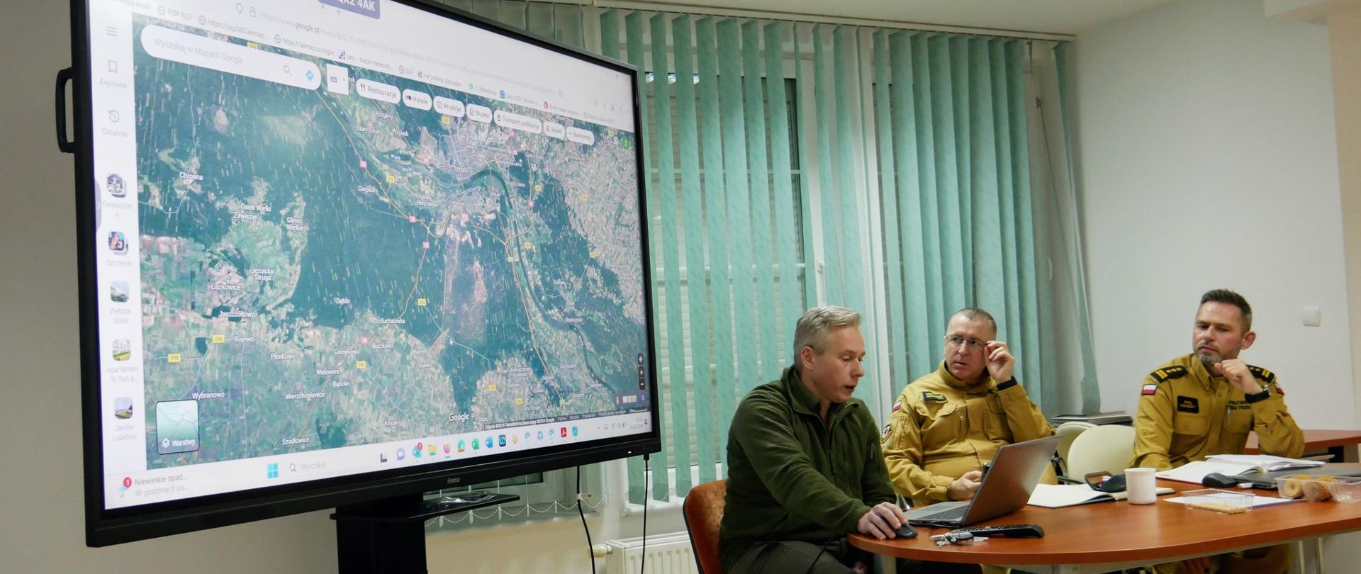 po prawej duży monitor konferencyjny na którym widać mapę terenu, po prawej stól prelegencki przy którym siedza zastępcy komendantów wojewódzkich i prelegujący lesnik