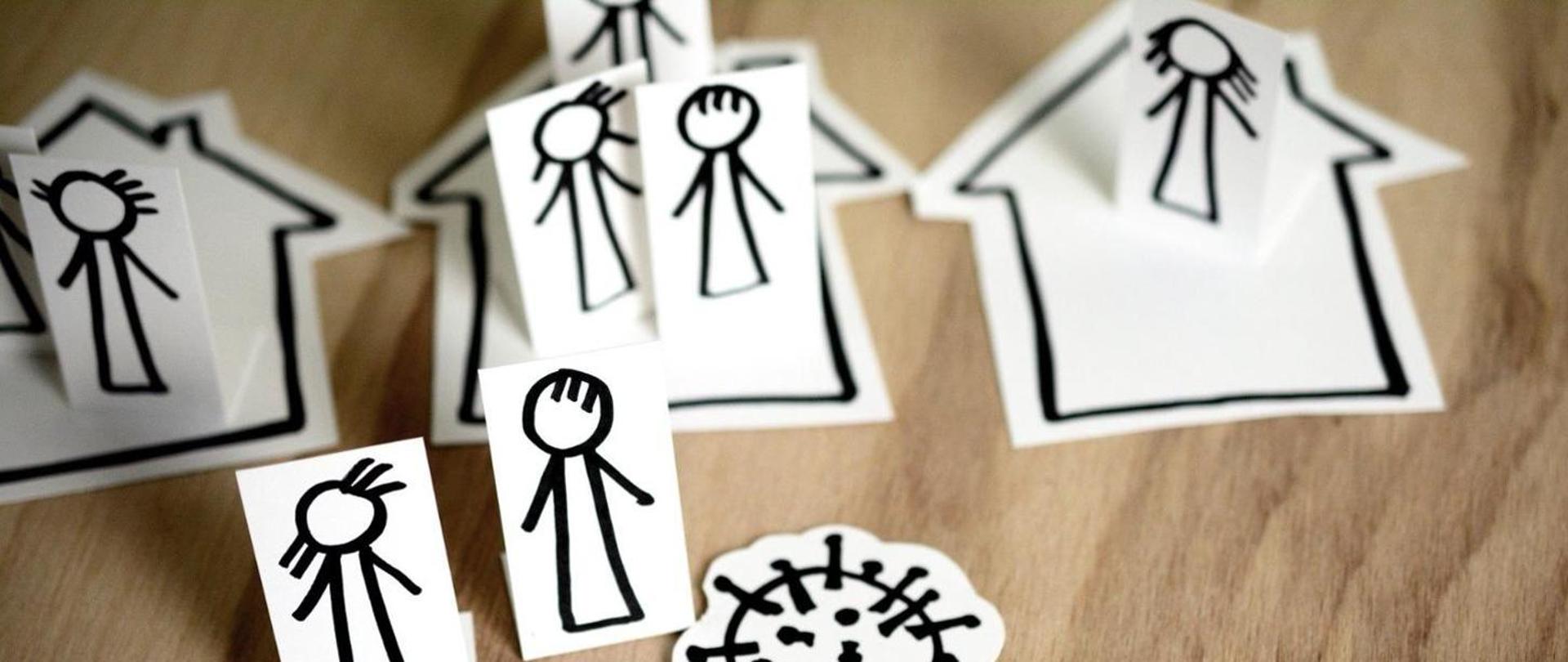 Białe karteczki z namalowanymi czarnym flamastrem postaciami ludzi. Karteczki stoją na drewnianym stole i są poustawiane w domach leżących na stole i poza nimi (domy są namalowane na kartkach, wyciętych w tym samym kształcie).