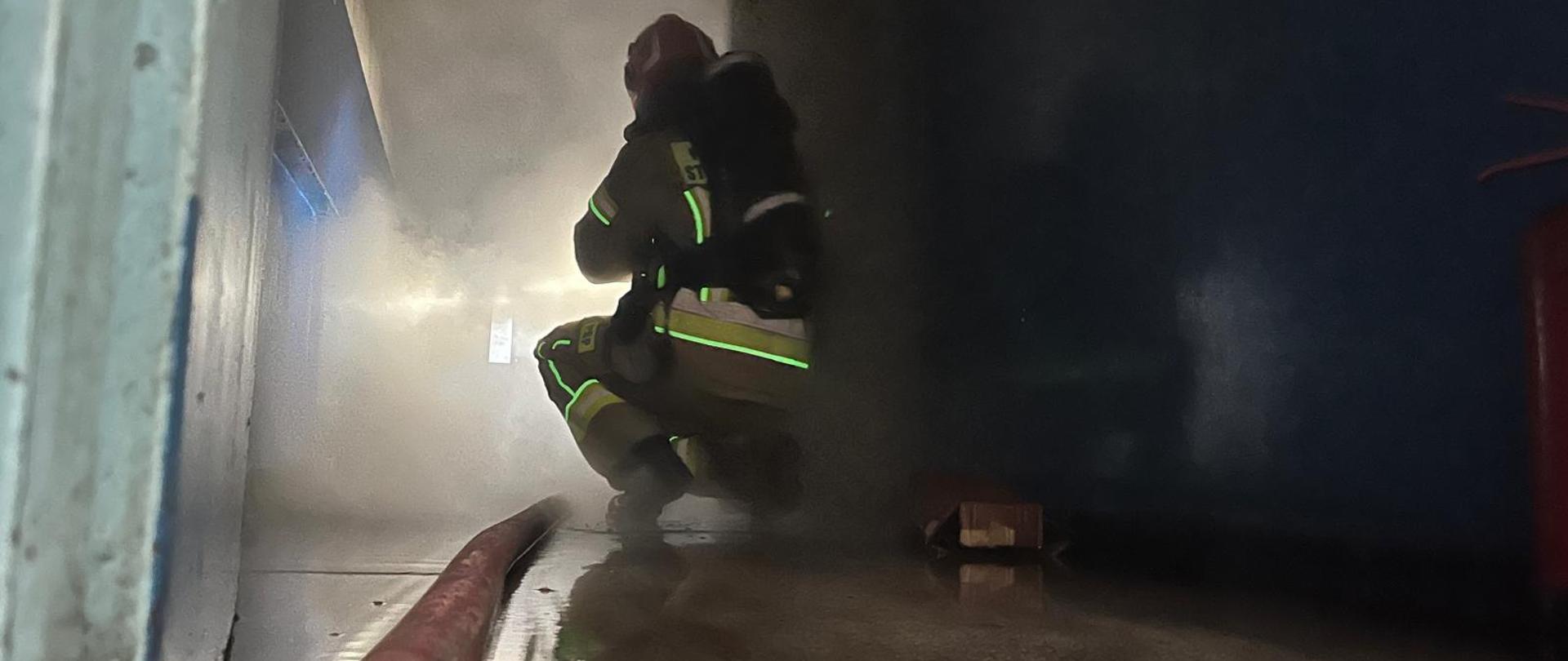 Ćwiczenia z gaszenia pożarów wewnętrznych. W tle duże zadymienie wydobywające się z pomieszczenia objętego pożarem, po prawej stronie strażak w pełnym umundurowaniu, w aparacie ochrony dróg oddechowych, na podłodze rozwinięta linia gaśnicza.