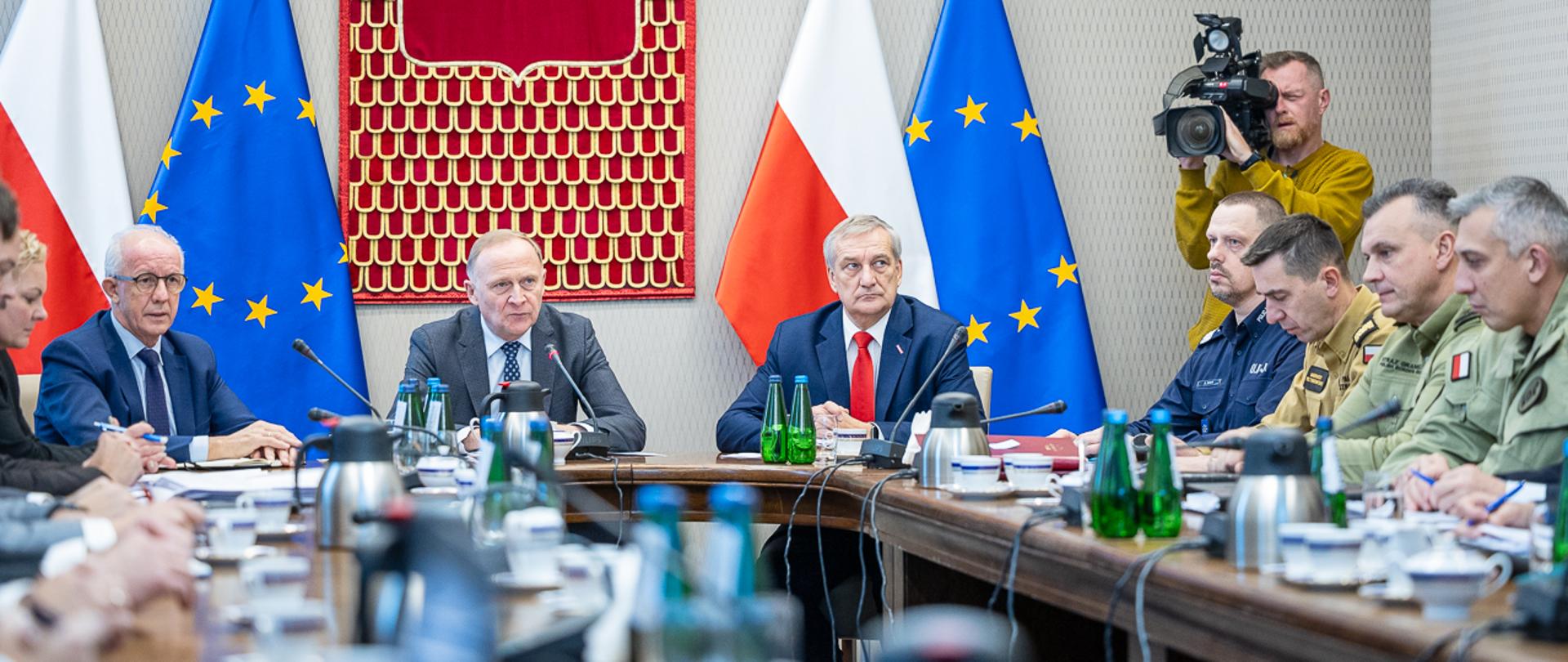 Siedmiu mężczyzn za stołem prezydialnym na tle flag Polski i UE. W prawej części kadru, operator kamery telewizyjnej. 