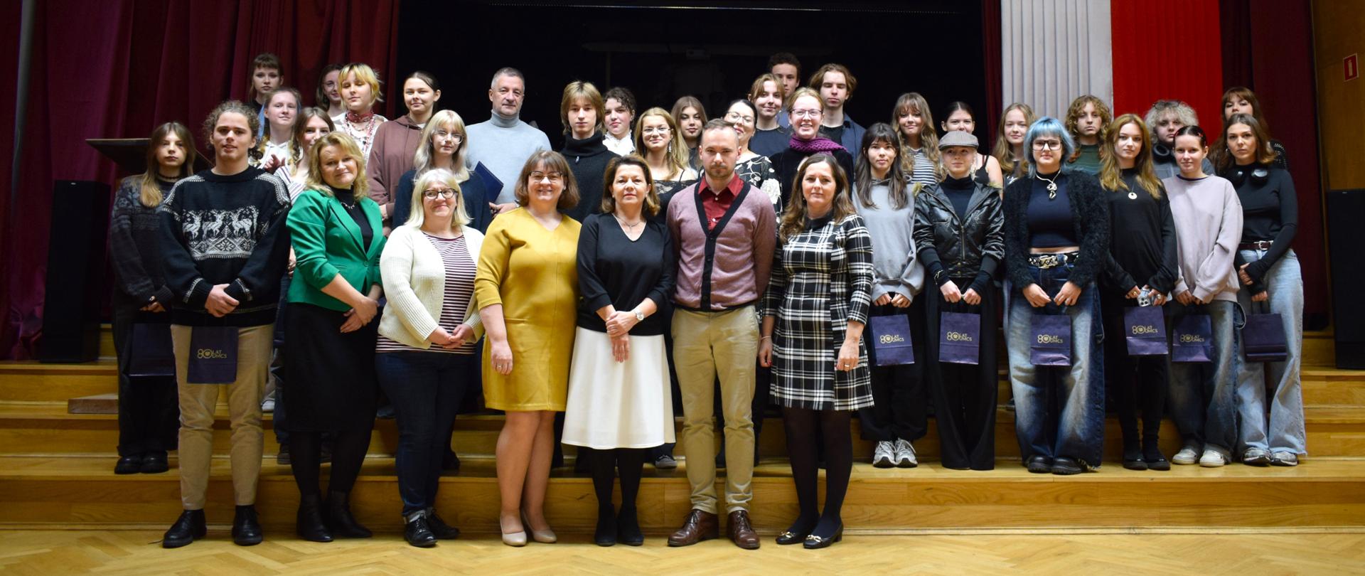 Grupa młodych oraz siedem dorosłych osób stoi w dużym pomieszczeniu (auli), na drewnianych schodach. Za nimi duże, czerwone kotary, flaga Polski, pod sufitem zawieszone godło Polski