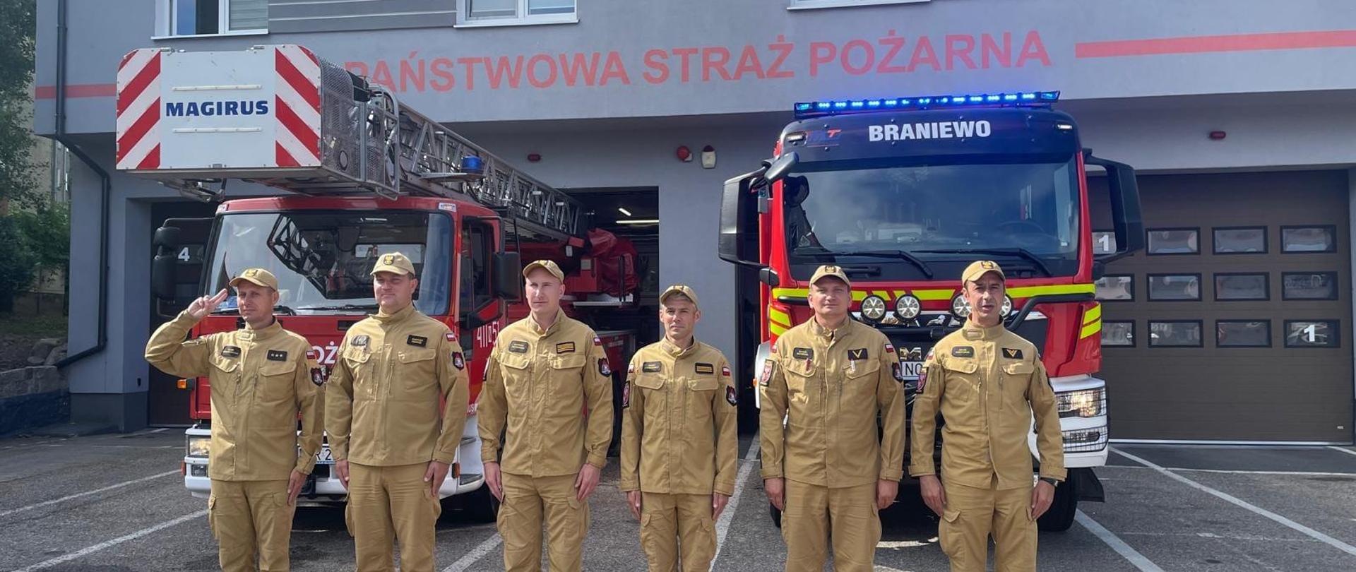 Sześciu strażaków w jasnych mundurach na tle samochodów strażackich i strażnicy. 