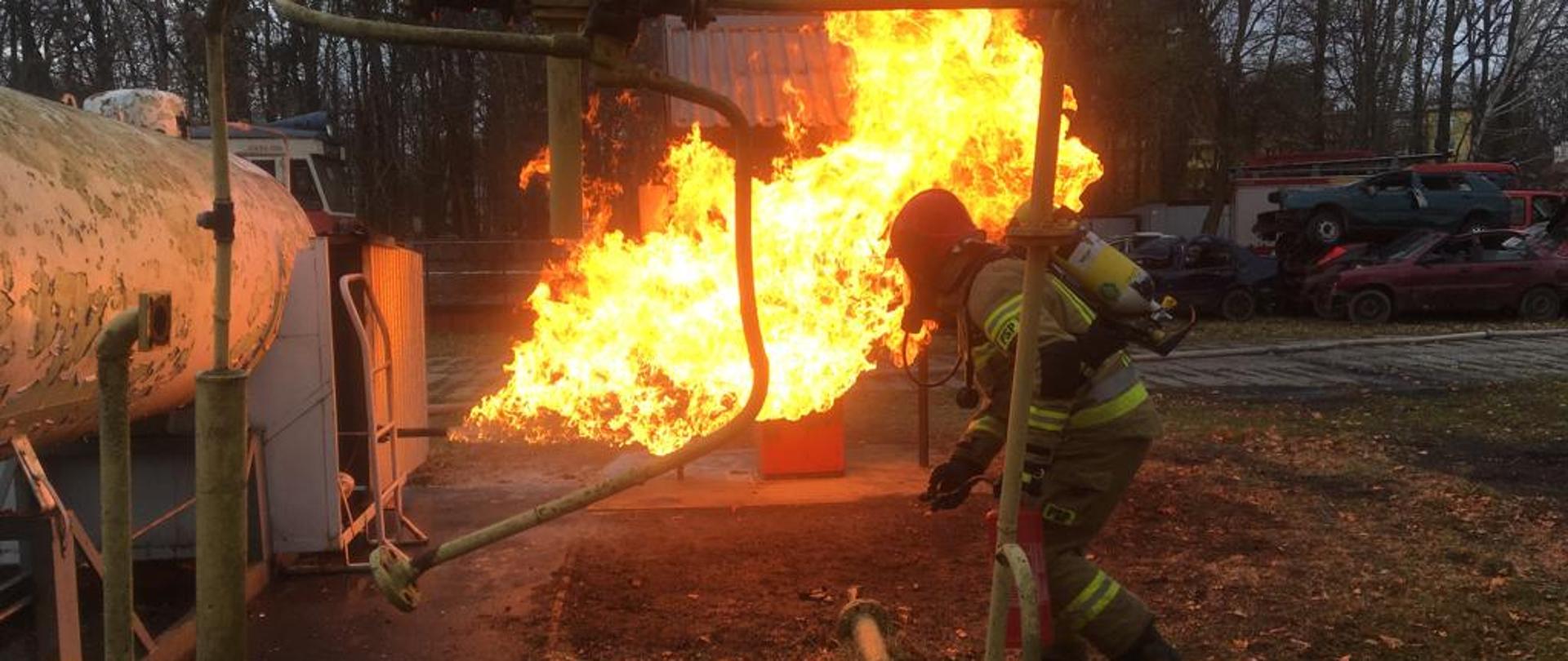 funkcjonariusz PSP podczas zajęć na poligonie szkoleniowym Ośrodka Szkolenia w Pionkach KW PSP w Warszawie gasi rozszczelniony zbiornik instalacji LPG. Ubrany jest w aparat ochrony dróg oddechowych oraz ubranie specjalne.