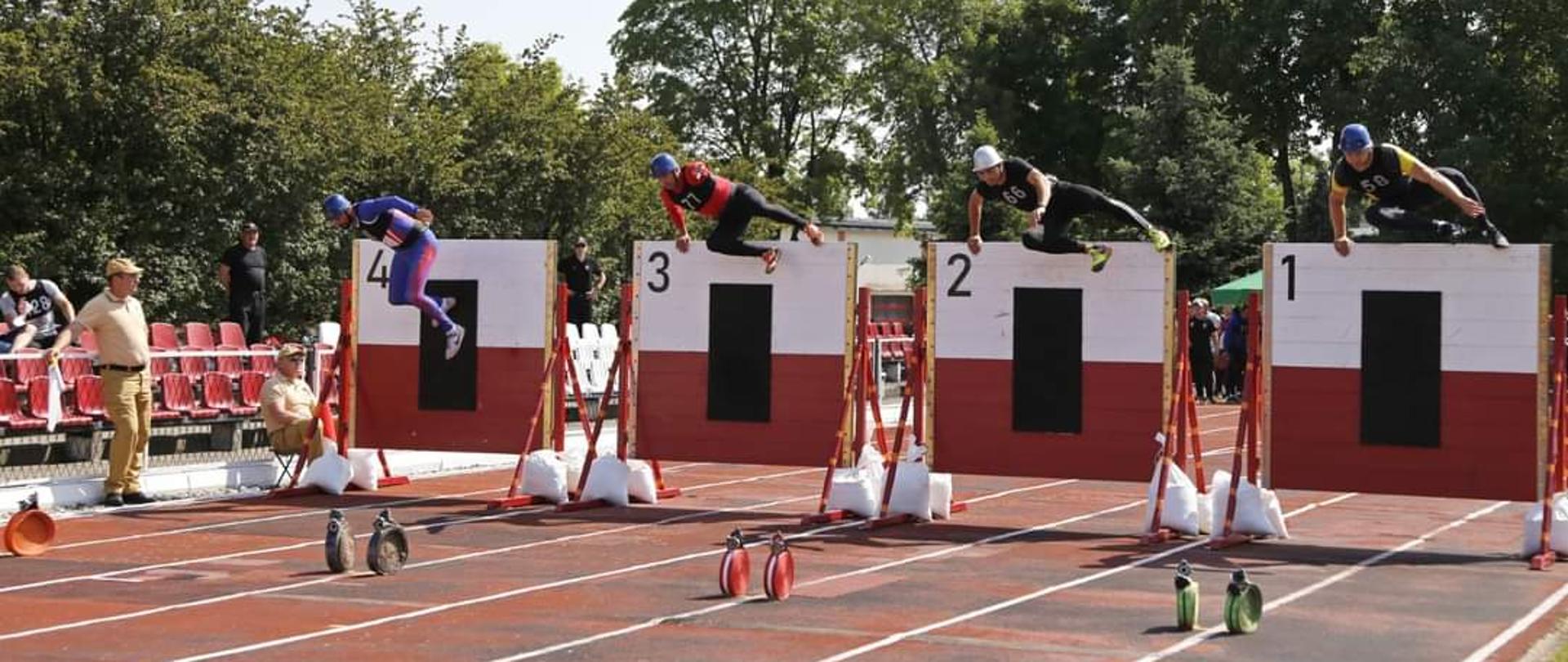 Zdjęcie przedstawia czterech zawodników pokonujących dwumetrową ścianę w konkurencji pożarniczy tor przeszkód 100 metrów.