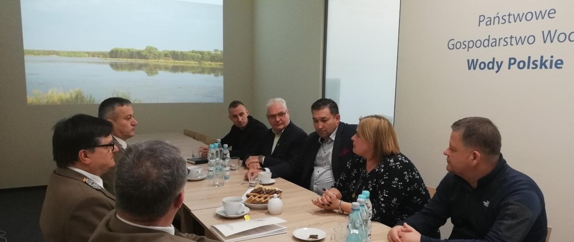 Jasny pokój z dużym stołem. Przy stole siedzą uczestnicy spotkania. Na jednej ścianie widoczne logo Wód Polskich, na drugiej wyświetlana jest prezentacja z rzutnika.