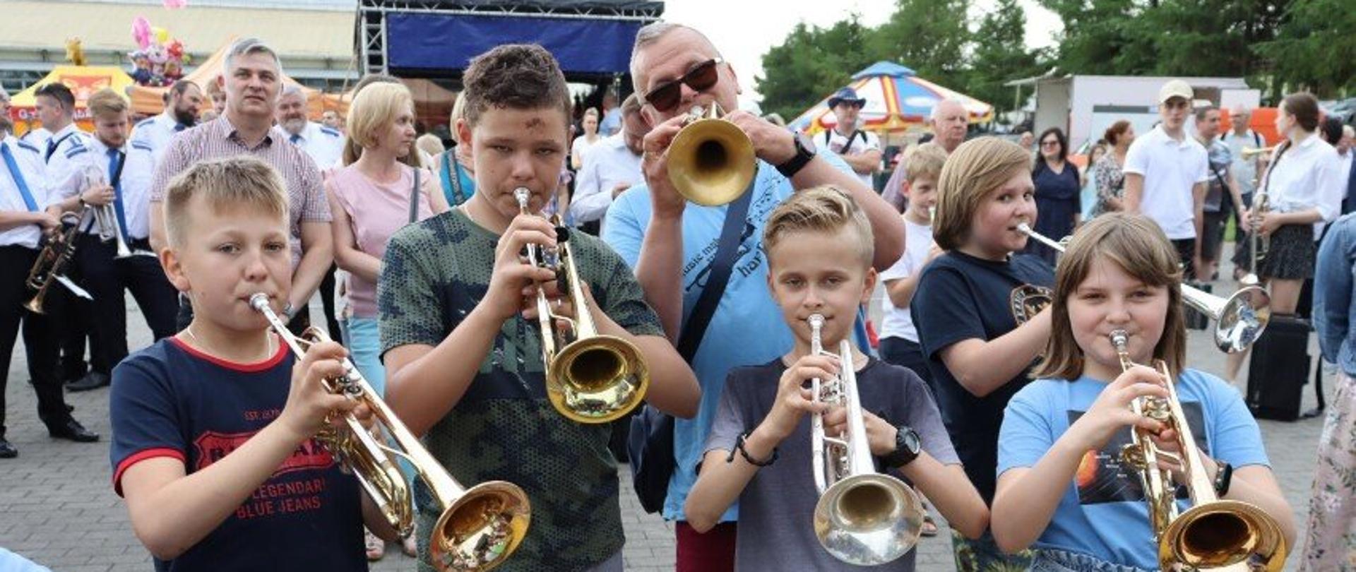 Zdjęcie przedstawia pięciu uczestników konkursu wraz z opiekunem. Trzymają przy ustach instrumenty. W tle widać niewyraźne sylwetki innych uczestników konkursu.