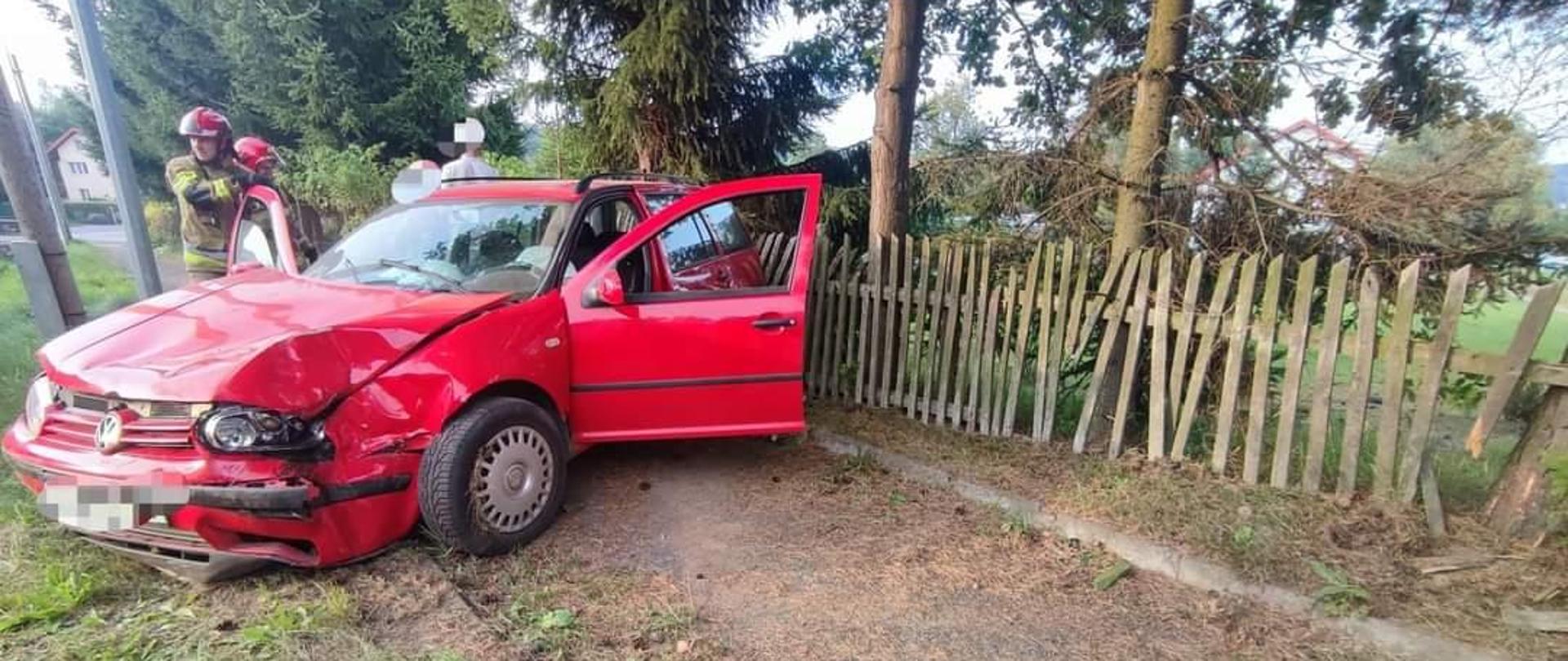 Samochód osobowy marki Volkswagen Golf z uszkodzonym przodem. Auto w kolorze czerwonym, za autem dostrzegamy strażaka w ubraniu specjalnym.