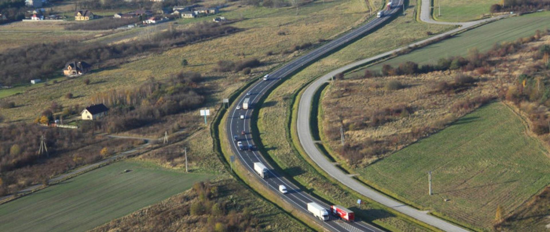 Widok z lotu ptaka na pojazdy jadące po łuku drogi. Po bokach drogi widoczne pola i nieliczne zabudowania.