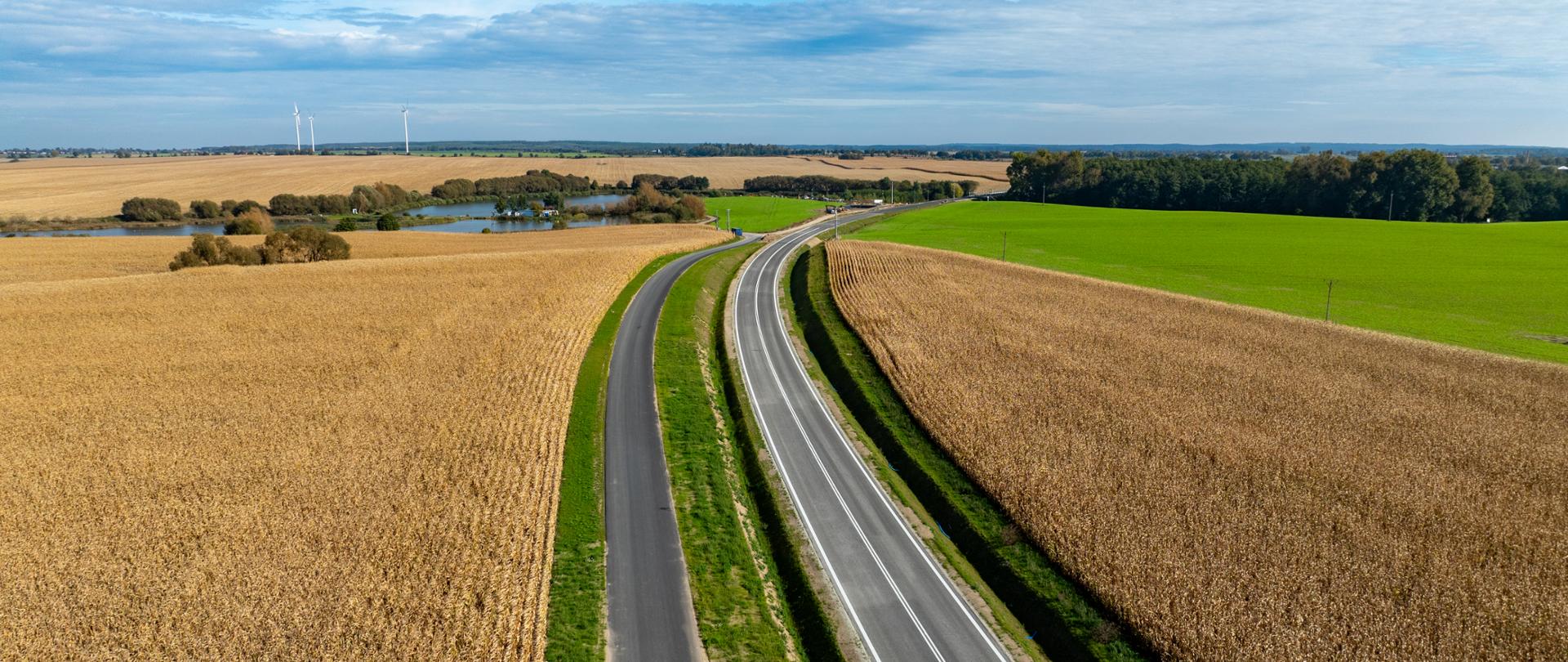 Obwodnica Strzelec Krajeńskich wijąca się między polami z dojrzałym zbożem. W oddali widać zbiornik wodny - staw oraz fragmenty lasu.