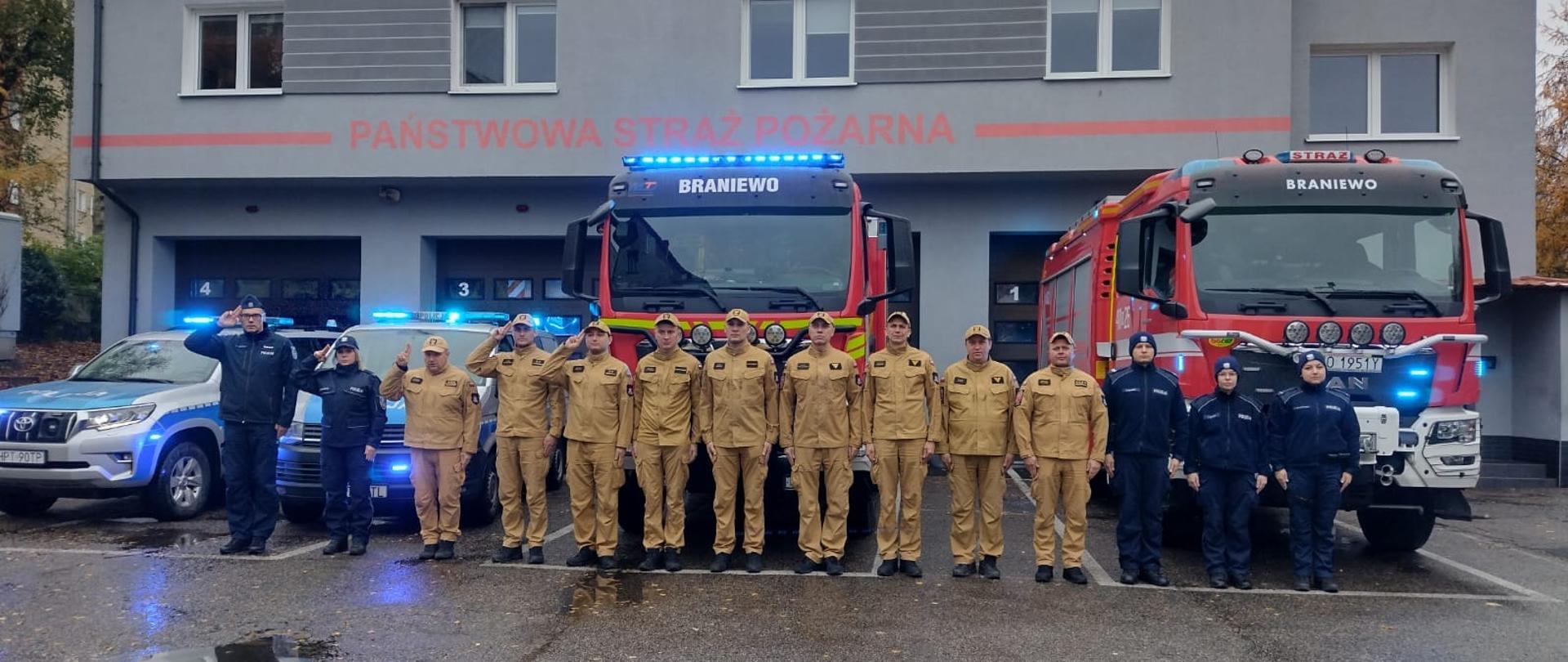 Na zdjęciu w szeregu ustawieni policjanci i strażacy. W tle samochody strażackie i z lewej strony dwa radiowozy.