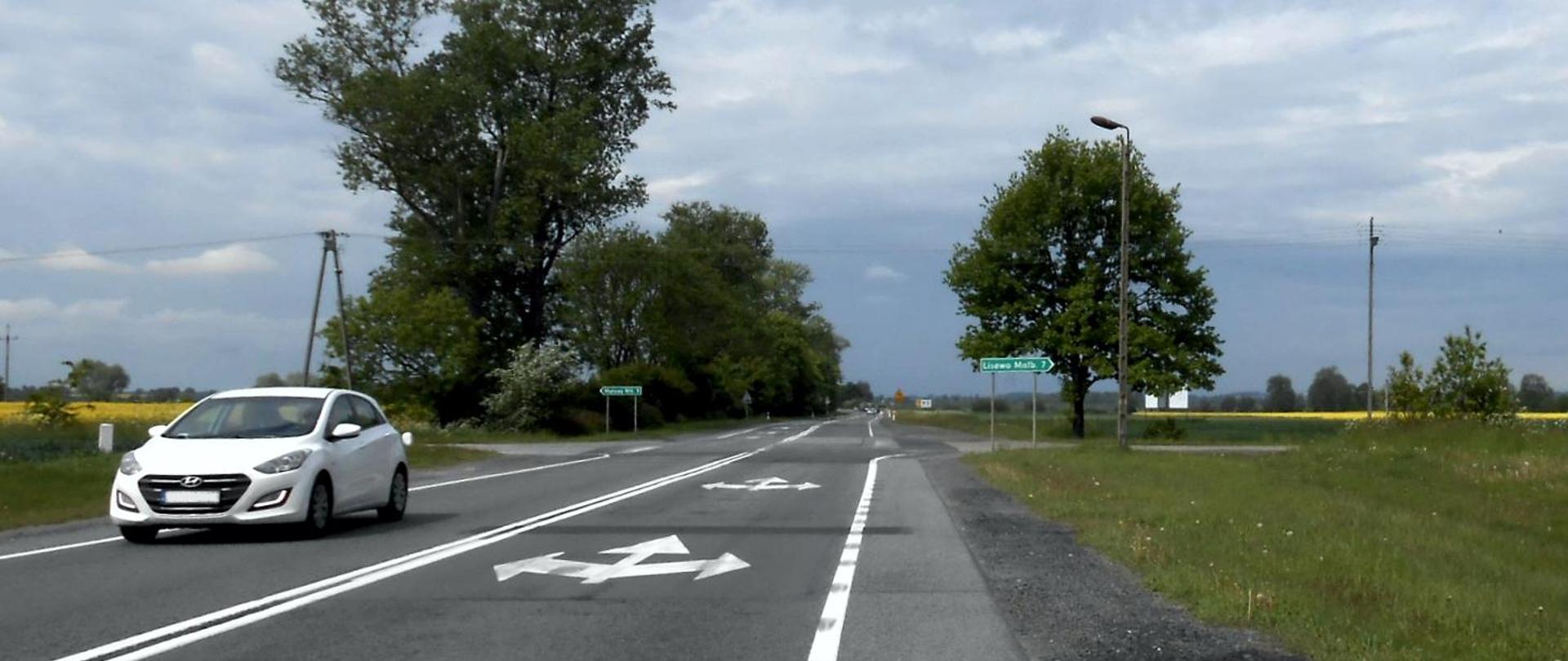 Letni dzień. Biały samochód jedzie drogą krajową w otoczeniu drzew i pól.